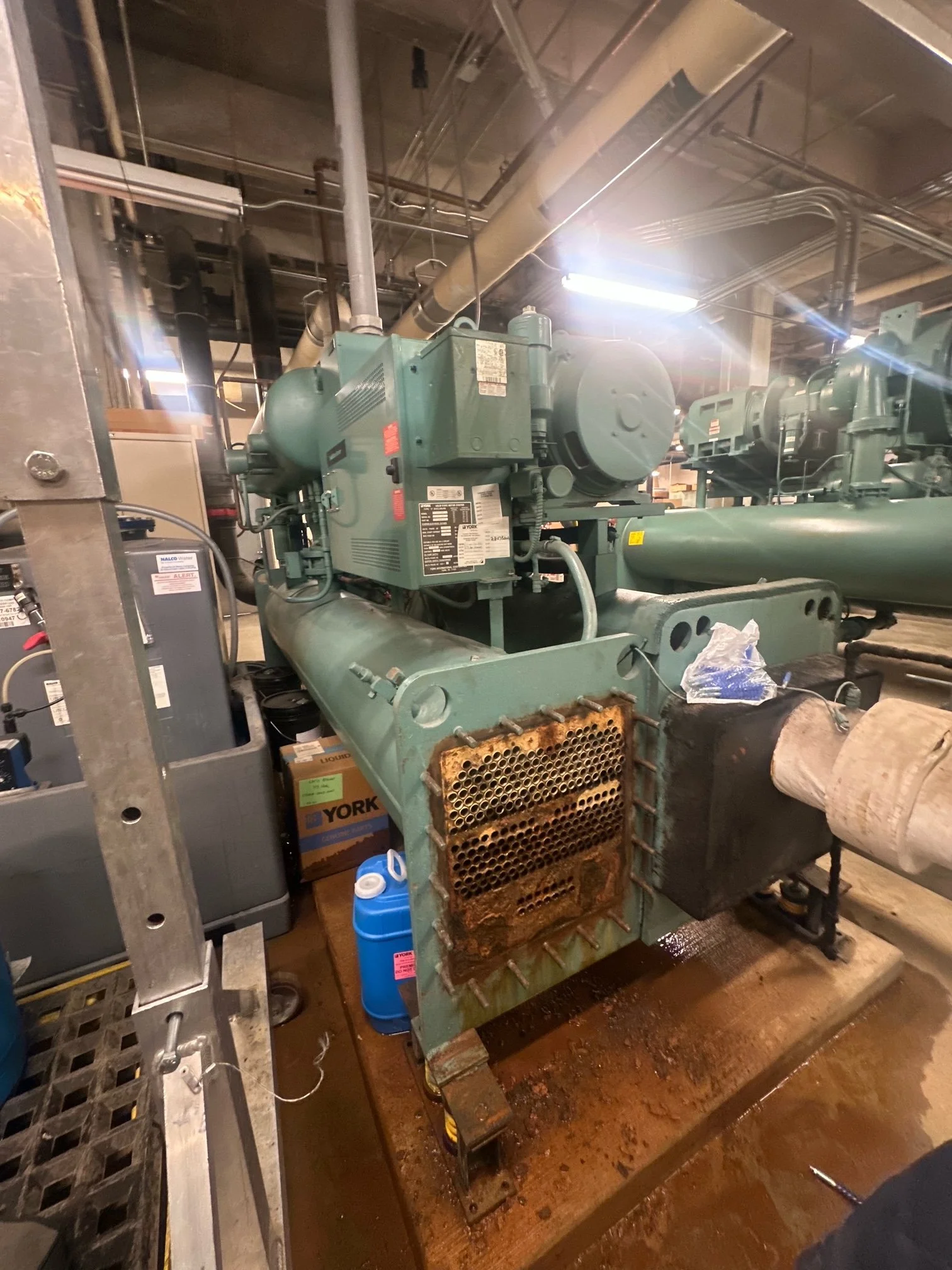 Industrial machinery with pipes and components in a factory setting, displaying a corroded section on the front.