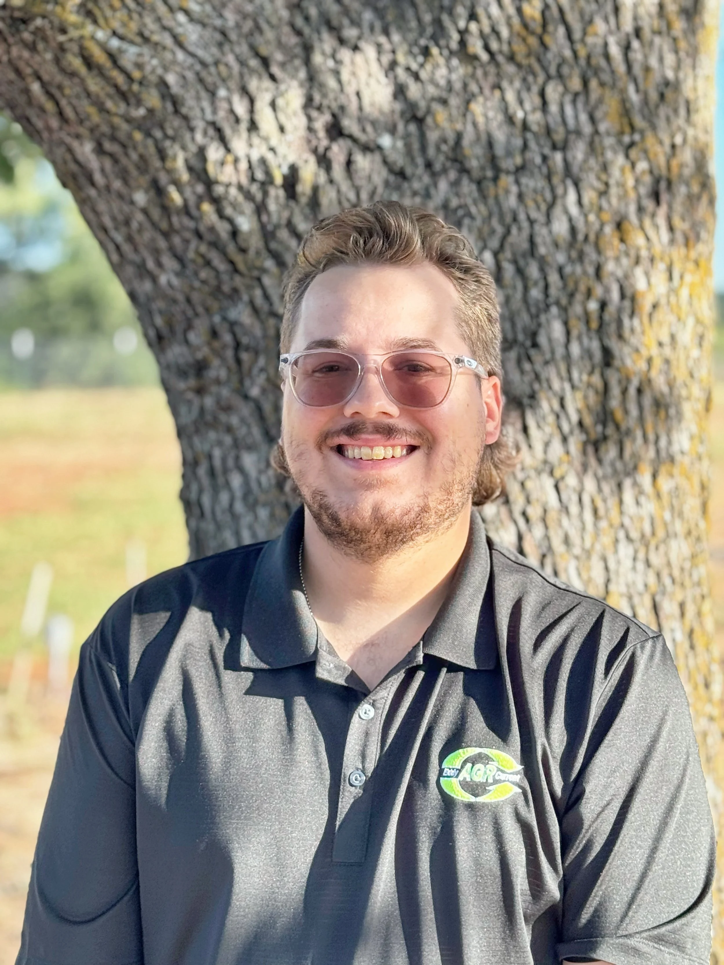 Smiling man with glasses wearing a black polo shirt with a logo, standing outdoors in front of a large tree trunk.