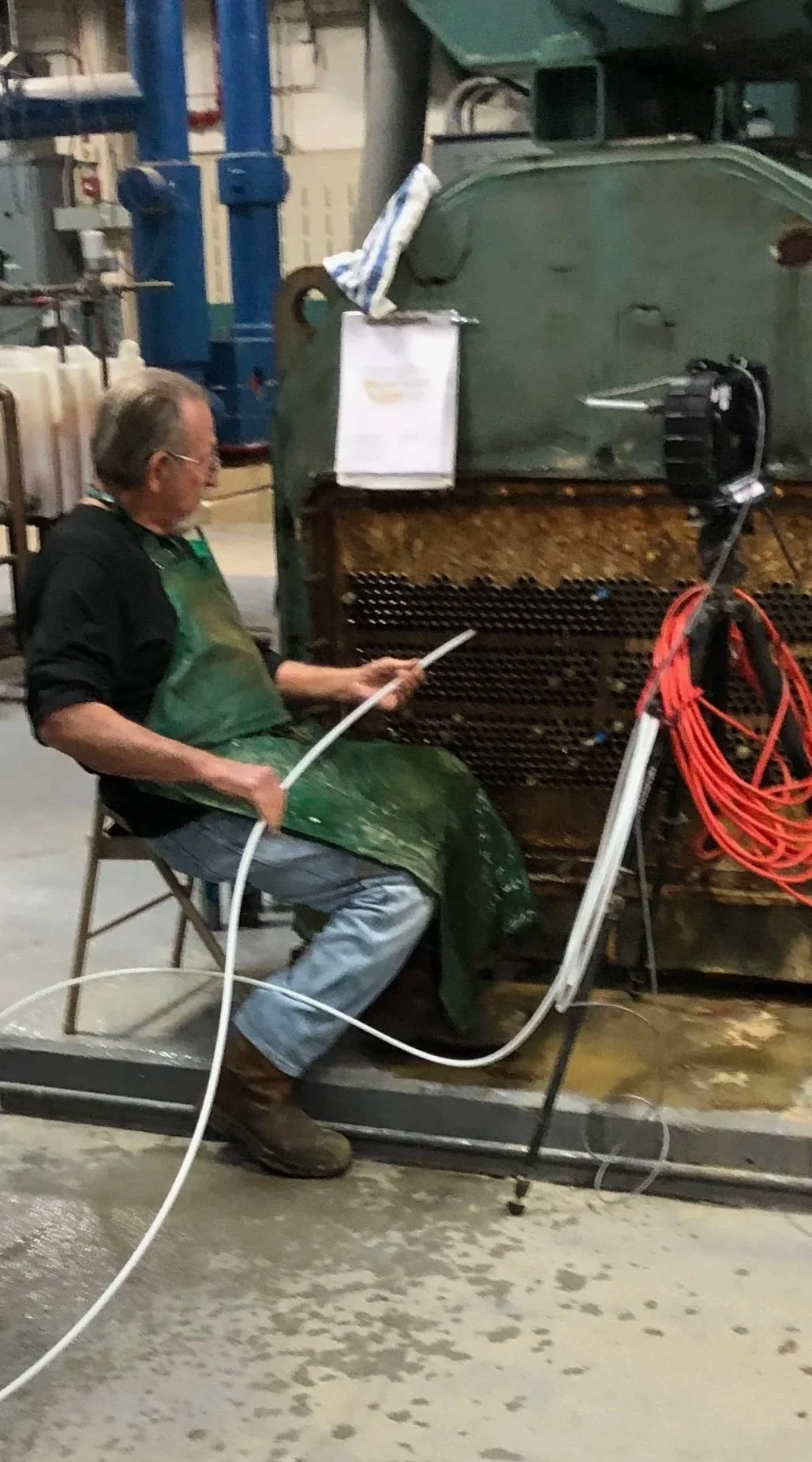 A man wearing a green apron and brown boots sitting on a chair in an industrial setting, working with a hose connected to a large machine with rust and grime, surrounded by industrial equipment and orange extension cords.