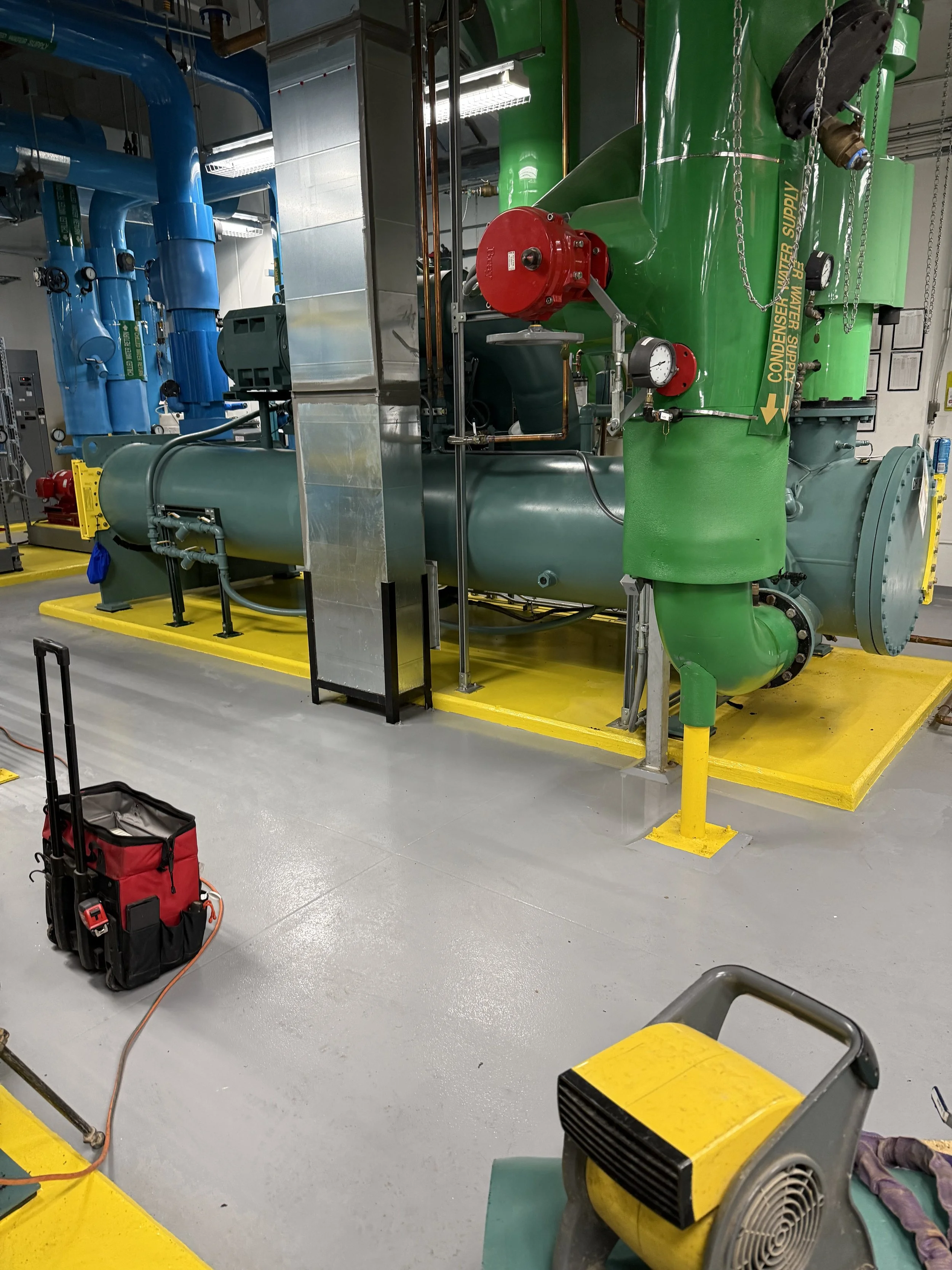 Industrial mechanical room with large green and blue machinery, yellow safety markings on the floor, tools, and equipment such as a red toolbox and a gray air blower, with pipes, gauges, and metal surfaces.