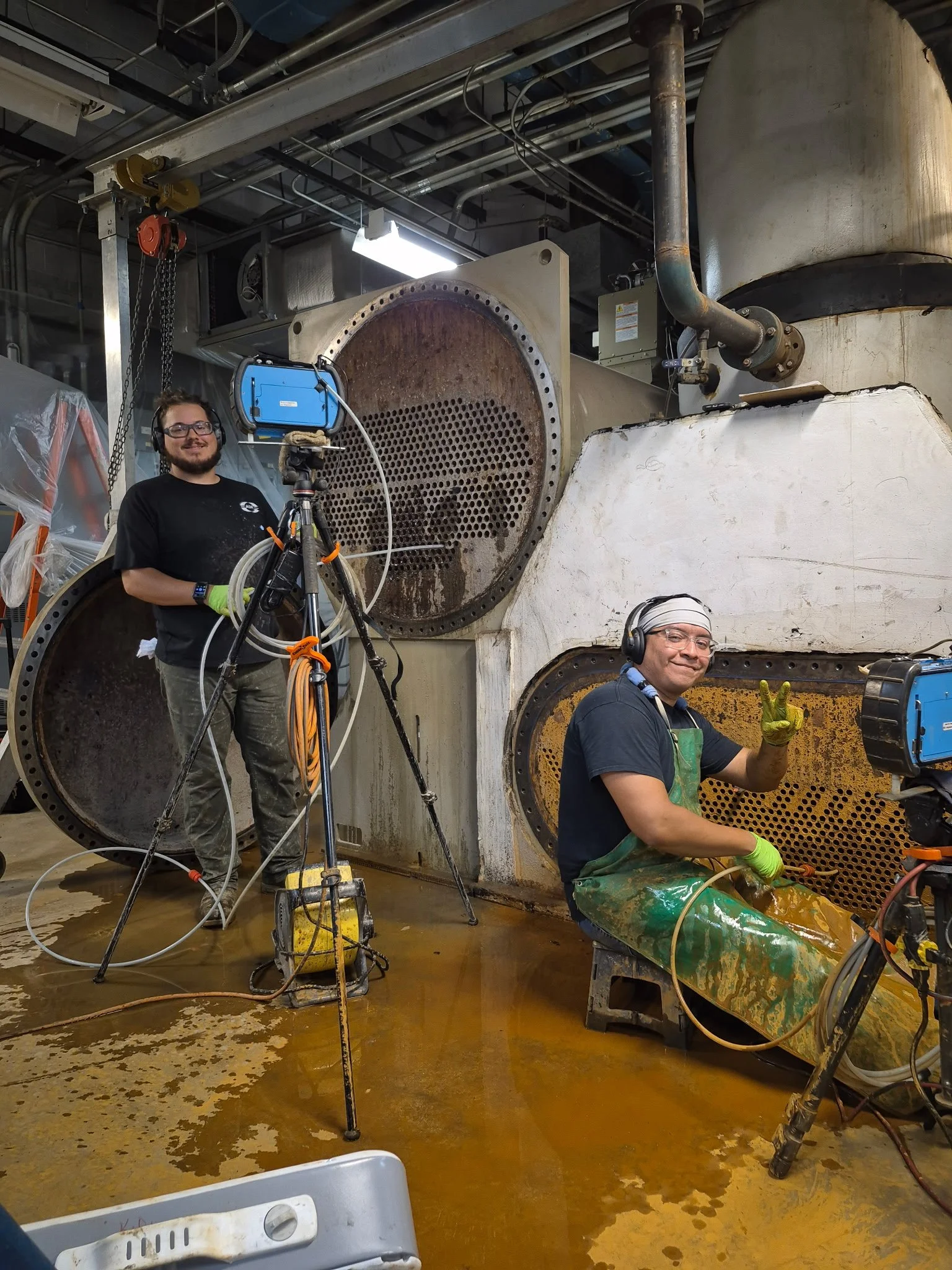 Two men working in a factory surrounded by large industrial equipment and pipes. They are wearing safety gloves, glasses, and hearing protection.