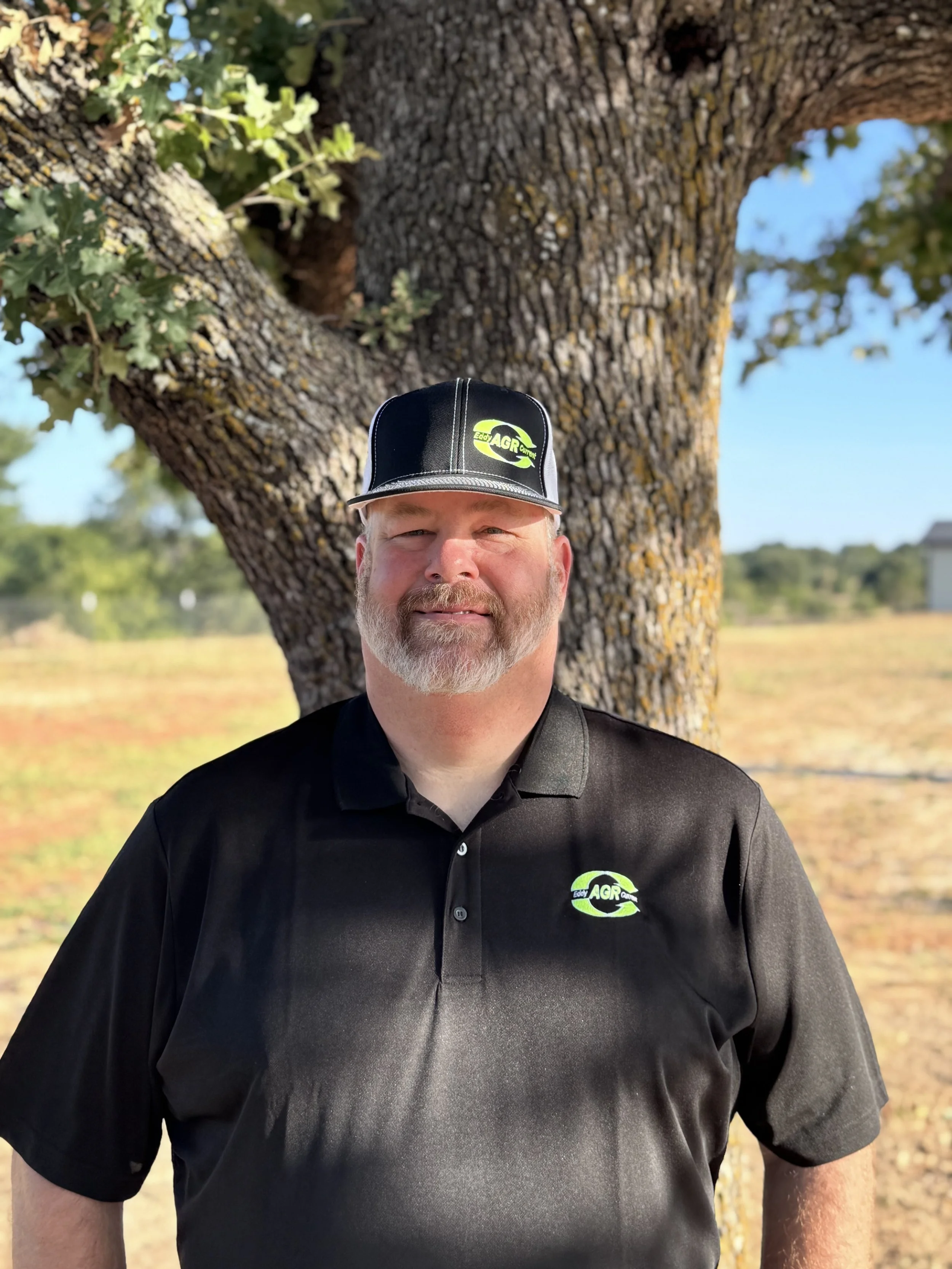 Man with white beard and mustache wearing a black shirt and a black cap with a green and white logo standing outdoors in front of a large tree with textured bark. The background is a rural landscape with clear skies.