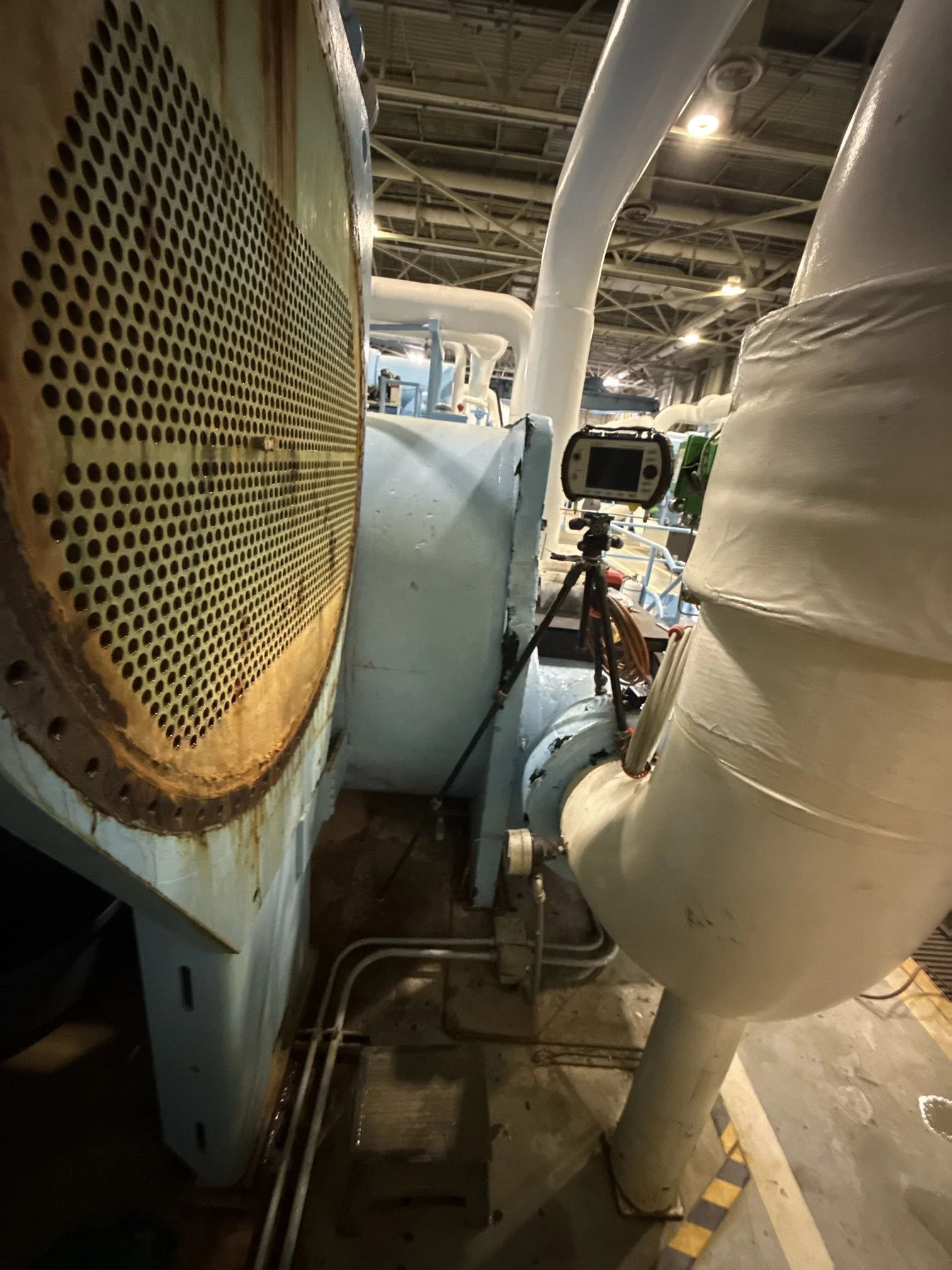 Industrial equipment with pipes, valves, and machinery in a factory setting, with a digital device mounted on a tripod in the foreground.