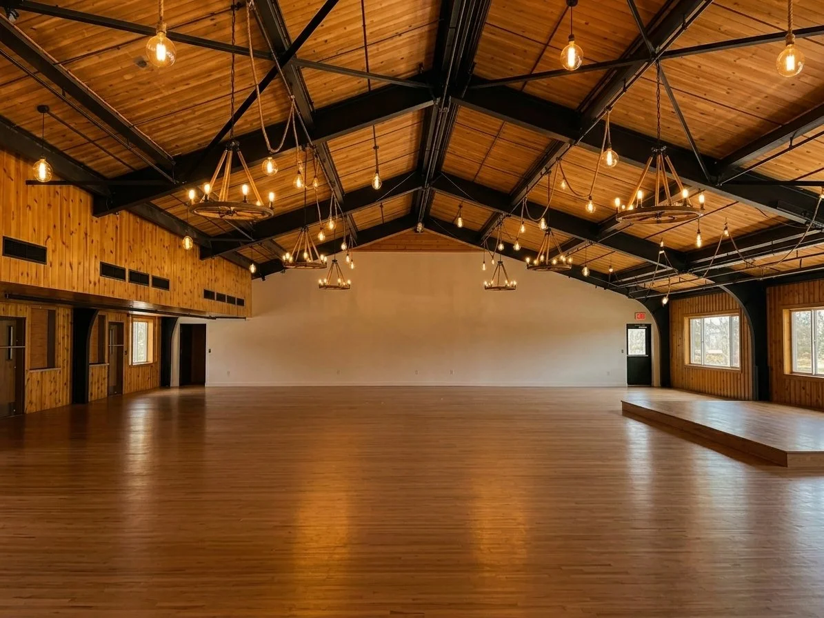 Empty rustic event hall with wooden floors, wood-paneled walls, and a wooden vaulted ceiling with exposed black beams and hanging Edison light bulbs. Large windows along the side wall provide natural light, and there is a small stage at one end of the room.
