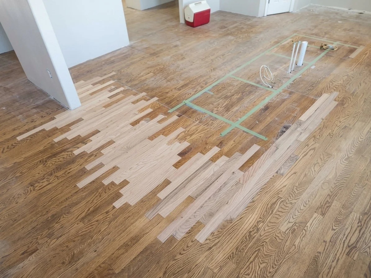 Partially installed hardwood flooring with sections of light and dark wood planks, some planks are laid down and others are stacked nearby, with tools and cables nearby.