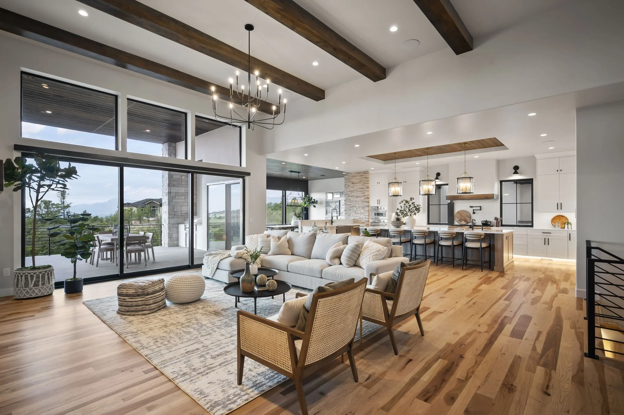 Open-concept living room with large windows, white walls, wooden ceiling beams, a chandelier, a beige sectional sofa with throw pillows, a round coffee table, and decorative poufs. The space extends to a dining area and kitchen, with patio doors lead
