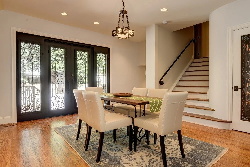 A dining area with a wooden table and six cream-colored chairs on a patterned rug, next to front glass doors with decorative ironwork, and a staircase with a black handrail in the background.