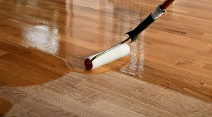 A paint roller with a handle is being used to apply a clear coat or sealant on a wooden floor.