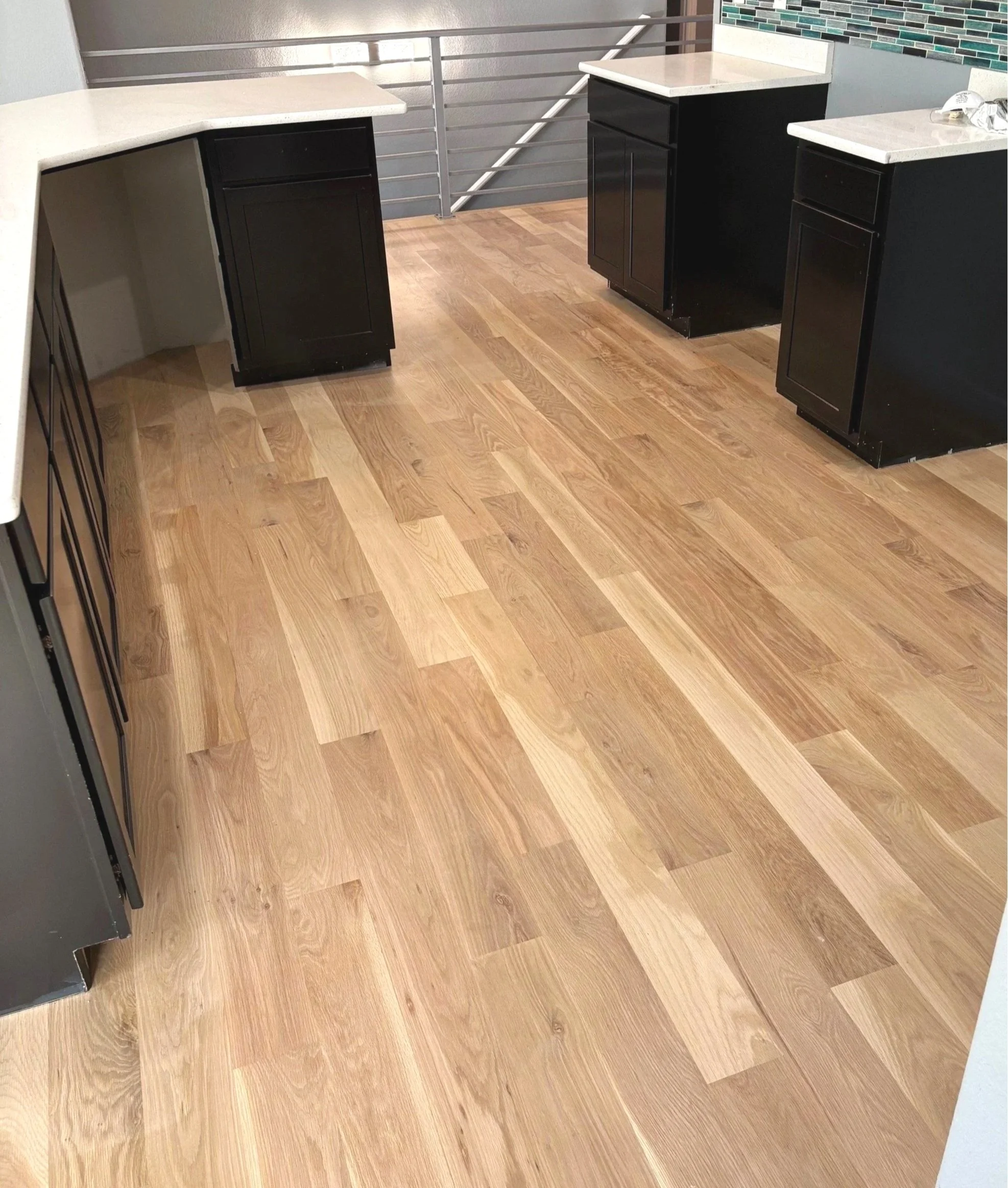 Interior view of a kitchen or dining area with wooden flooring, black cabinets, beige countertops, and a partial wall with mosaic tile accents.