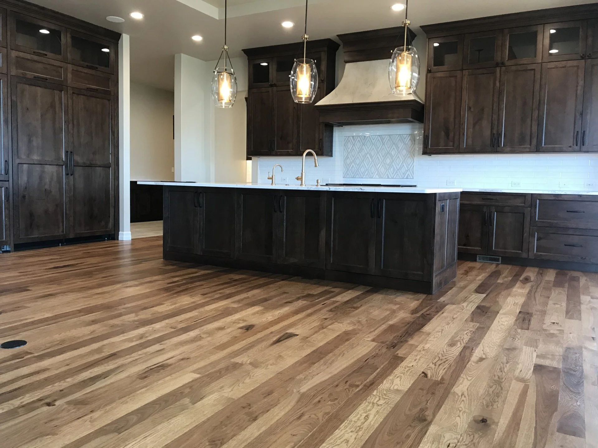 Modern kitchen with dark wood cabinets, white backsplash, and a kitchen island with a white countertop. Three pendant lights hang above the island, with wood flooring throughout.