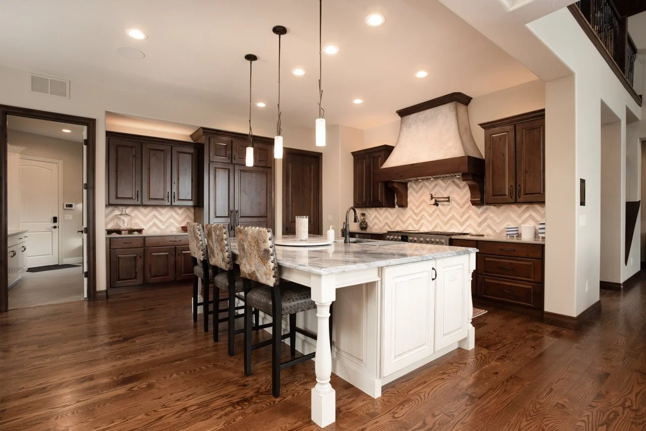 Modern kitchen with white island, dark wood cabinets, and chevron tile backsplash, pendant and recessed lighting, hardwood floors, and seating at the island.