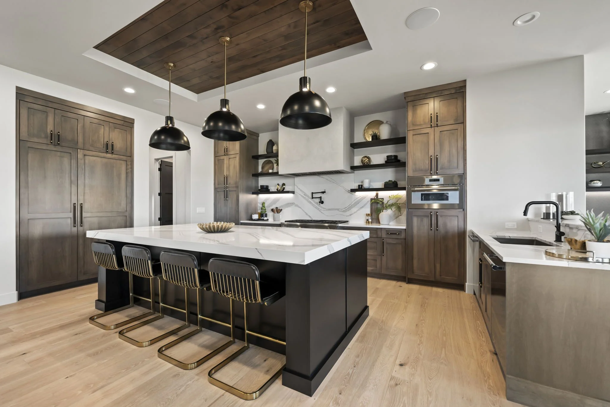 Modern kitchen with wood cabinetry, a large white marble island with black base, black pendant lights, open shelves, and a built-in oven.