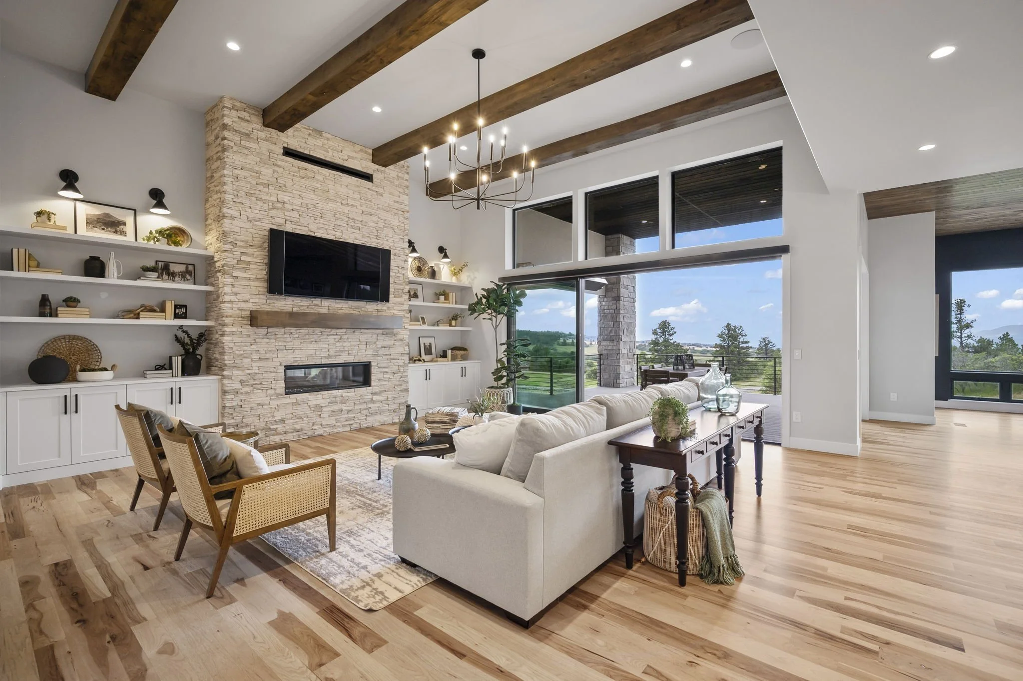 Living room with a stone fireplace, mounted flat-screen TV, white built-in shelves, large windows showing outdoor view, wooden ceiling beams, modern chandelier, beige sofa, wooden chairs, and decor.