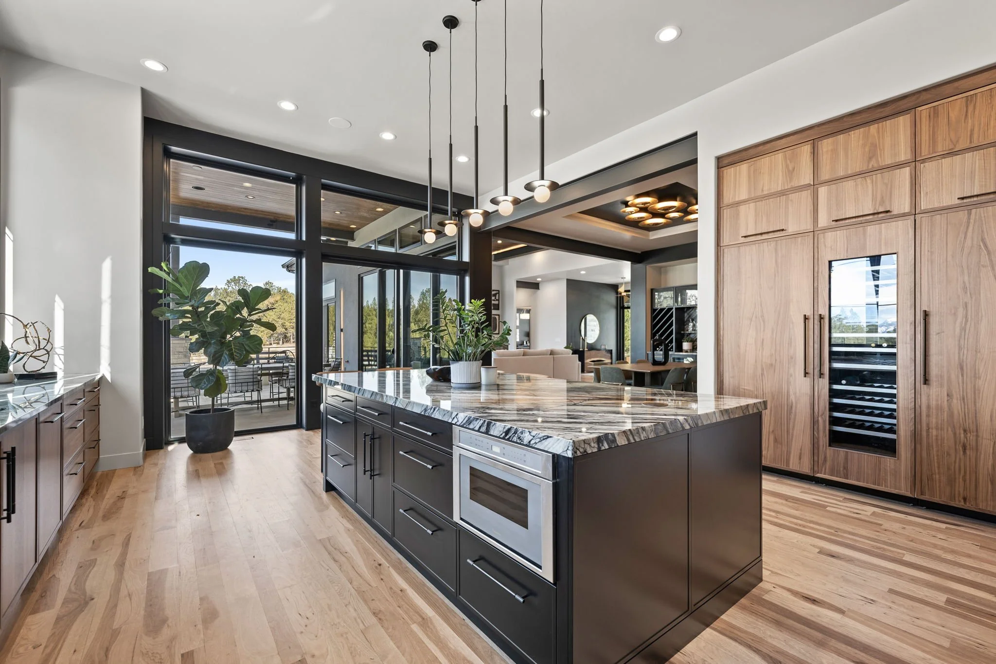 Modern kitchen with dark island, marble countertop, wooden cabinets, large glass sliding door, indoor plants, and open living space.