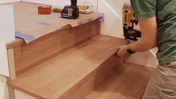 Person installing a wooden shelf or bench in a room, with tools and materials on the surface nearby.