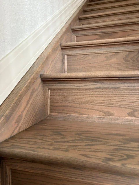 Close-up of wooden staircase with detailed crown molding and white baseboard against a white wall.