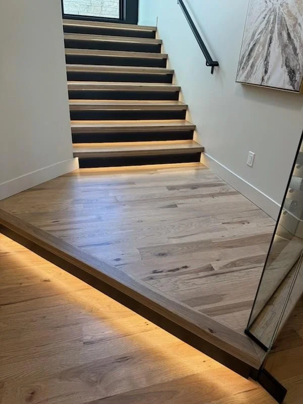 Modern staircase with black risers leading to a higher floor, illuminated by under-step lighting. The foreground shows a wooden floor with a reflective glass panel and a large abstract painting on the wall.