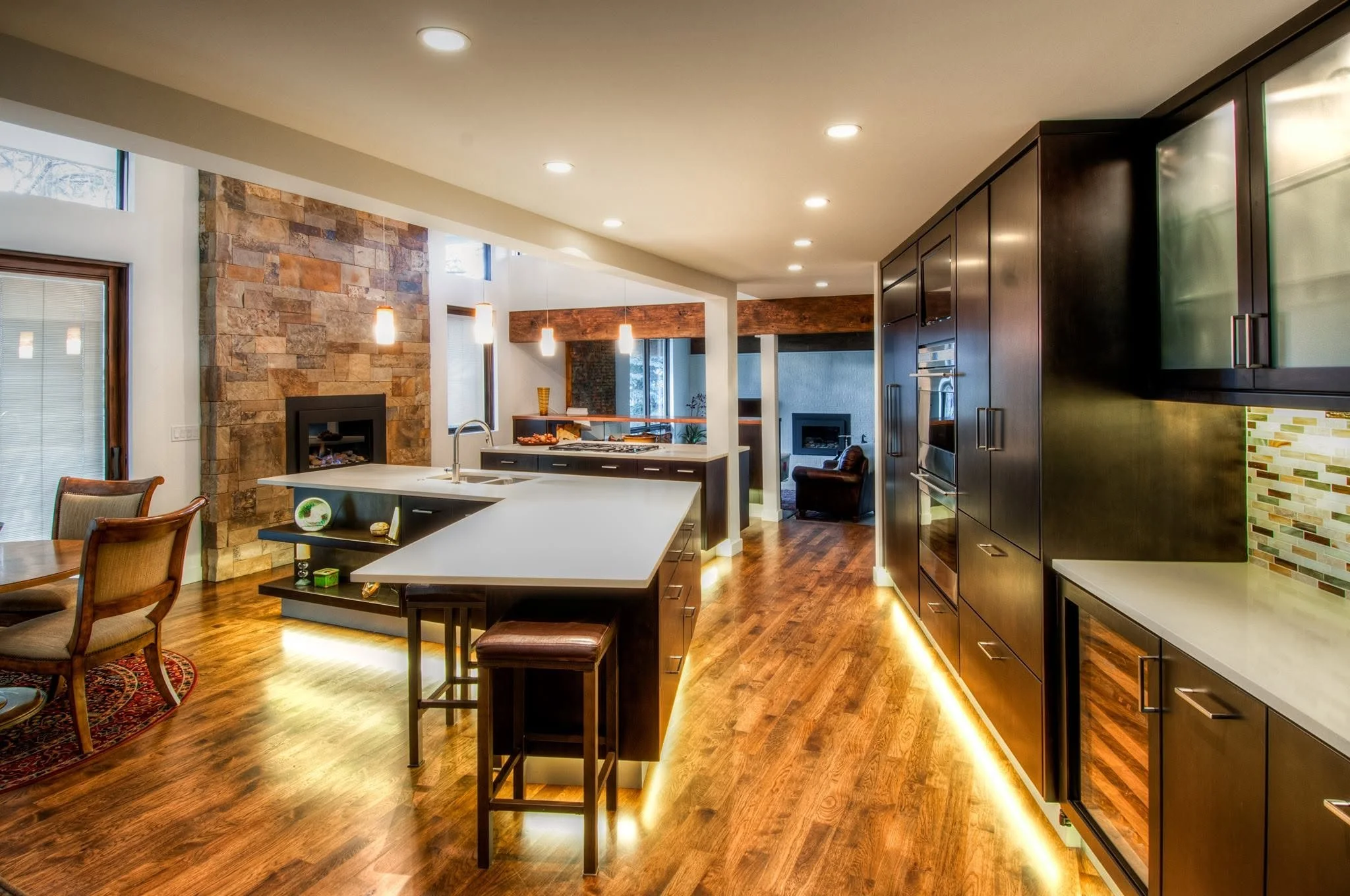 Modern open-concept kitchen with wooden floors, stone accent wall with fireplace, dark wooden cabinets, and a kitchen island with seating. Recessed lights and pendant lights create warm lighting.