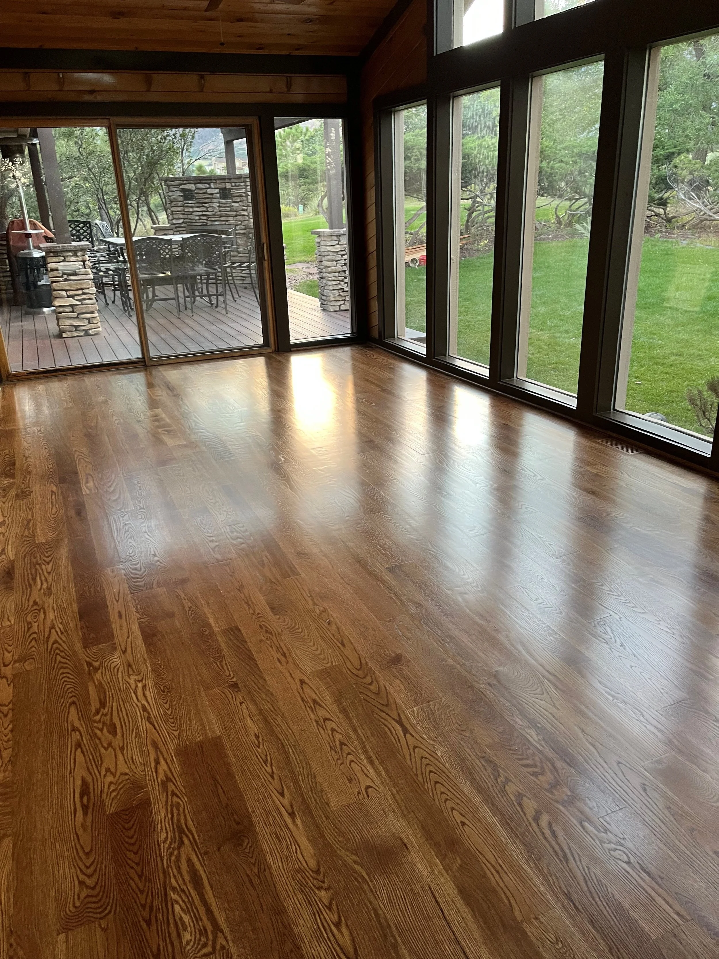Interior of a room with wooden floors and large windows overlooking a grassy yard and patio with outdoor furniture.