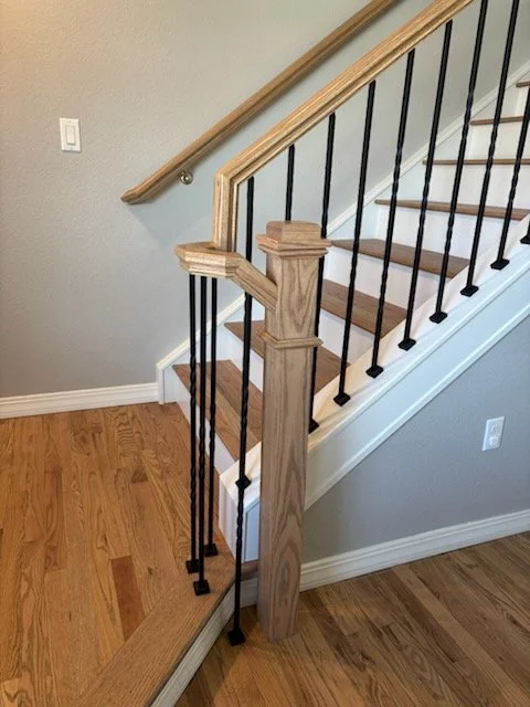 Interior staircase with wooden handrail and black metal balusters, hardwood stairs, and a gray wall with a light switch.