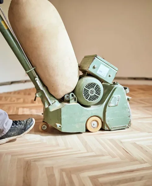 A green industrial floor polishing machine is cleaning a wooden floor, with a person's shoe visible nearby.