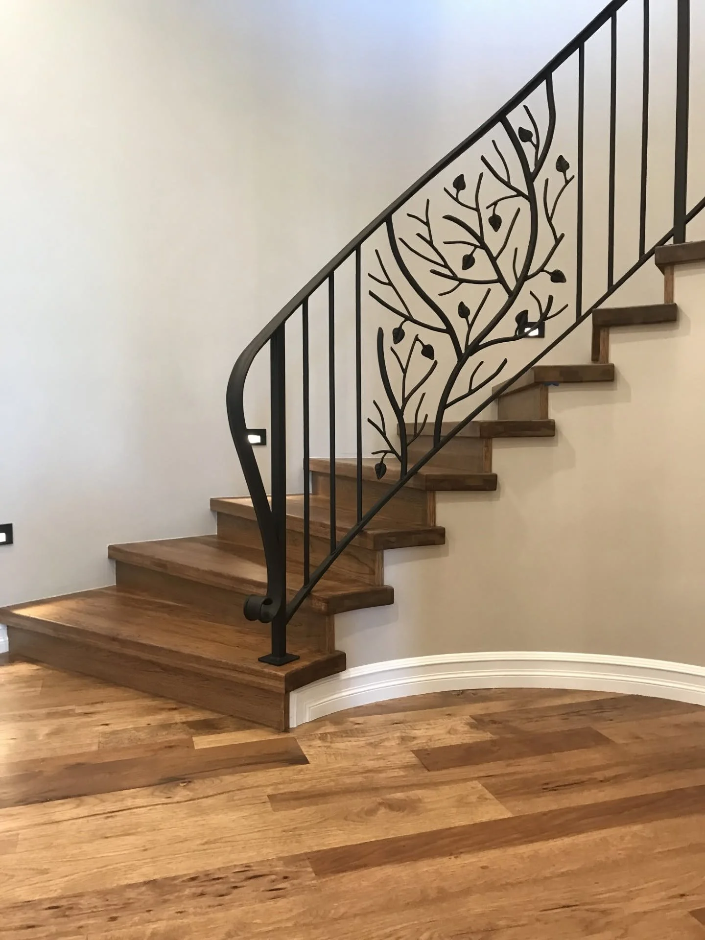Wooden staircase with decorative metal railing featuring a tree and leaf design, in a room with wooden flooring and white walls.
