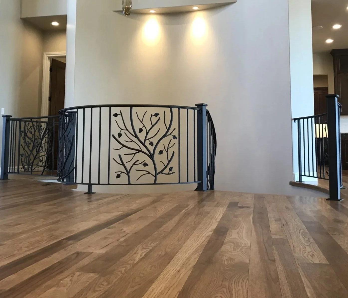 Interior view of a house with a curved black metal railing featuring a tree branch design, hardwood flooring, a beige wall with two wall-mounted lights, and a doorway leading to another room.