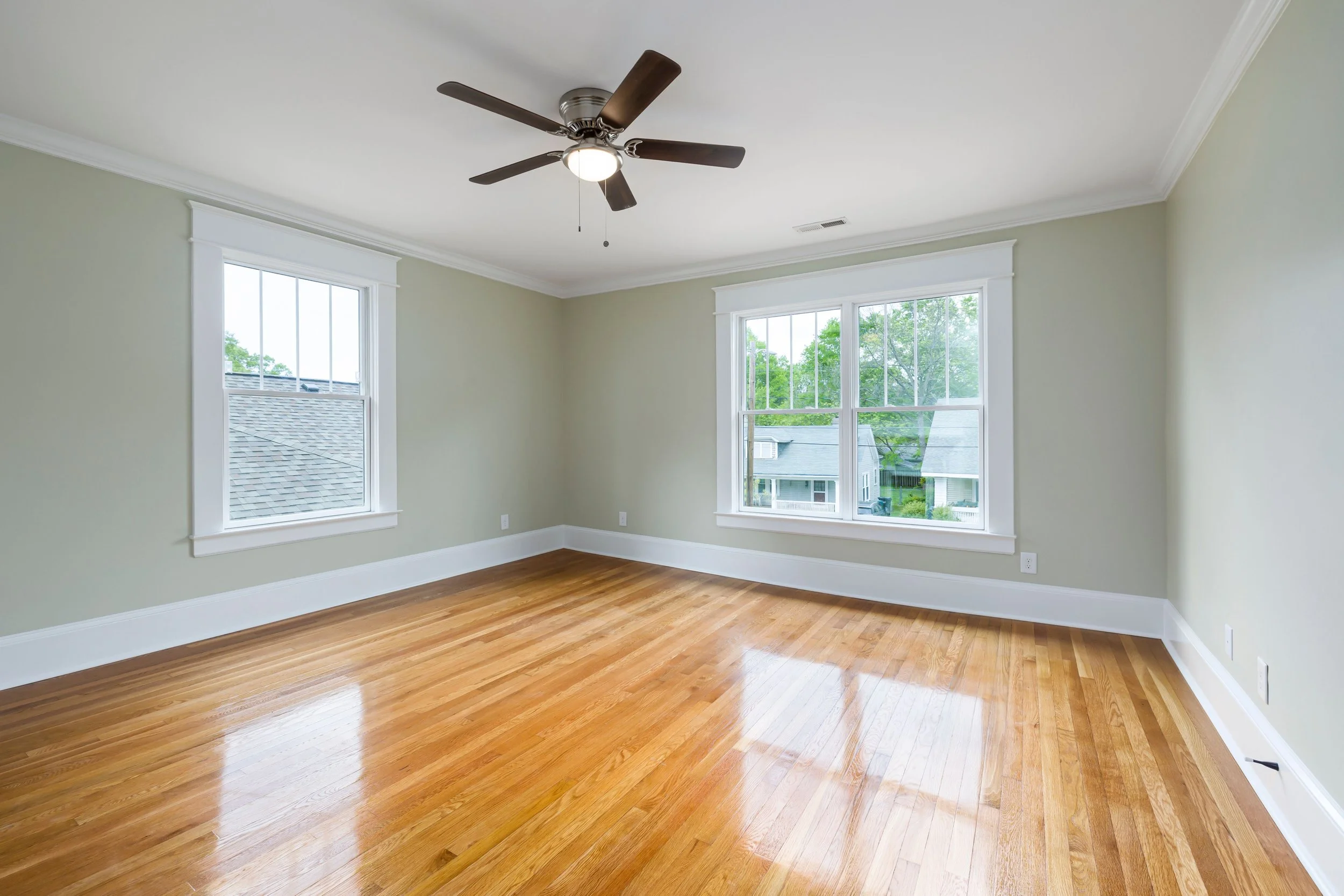 Empty room with hardwood floors, beige walls, white trim, two large windows showing houses outside, ceiling fan with light, and a vent in the ceiling.