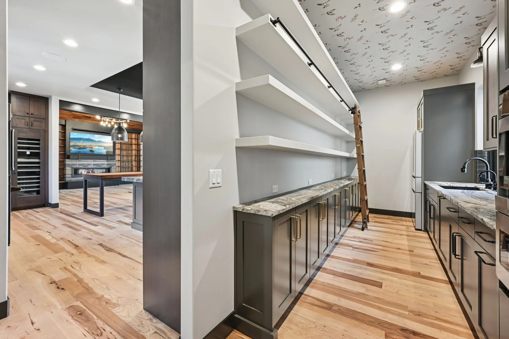 A modern open-concept kitchen with grey cabinetry, granite countertops, and stainless steel appliances. There's a kitchen island with a marble countertop and a ladder leaning against open white shelves. The ceiling has a unique pattern, and the room 