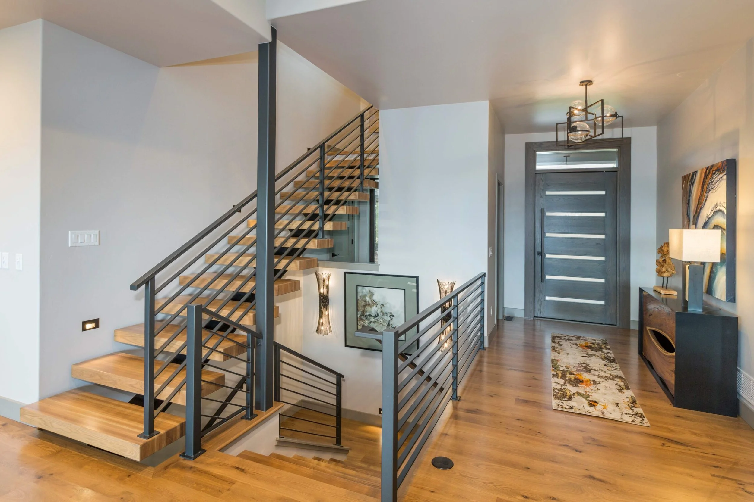 Interior of a modern home with stairs, front door, artwork, and furniture. Wooden flooring, metal stair railing, and decor accents.