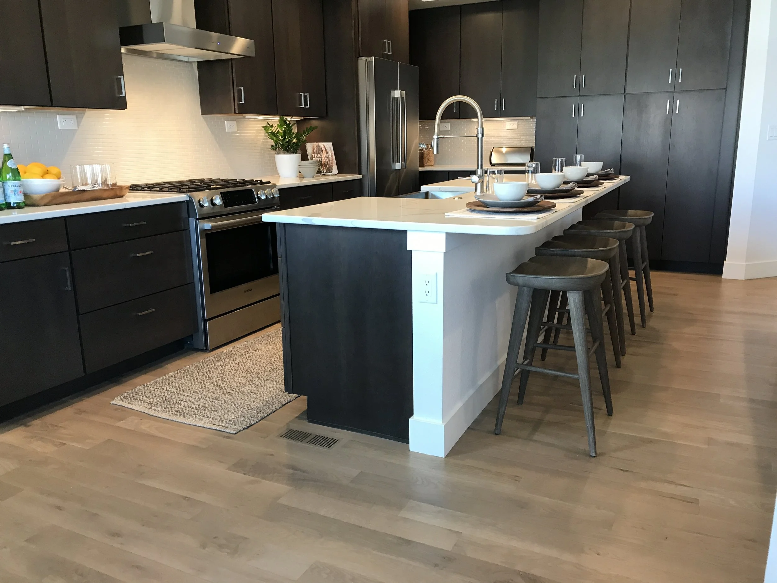 Modern kitchen with dark cabinets, stainless steel refrigerator, stove, and hood. White countertops, a small island with place settings, and bar stools. Decor includes a potted plant, a bowl of lemons, and a cookbook.