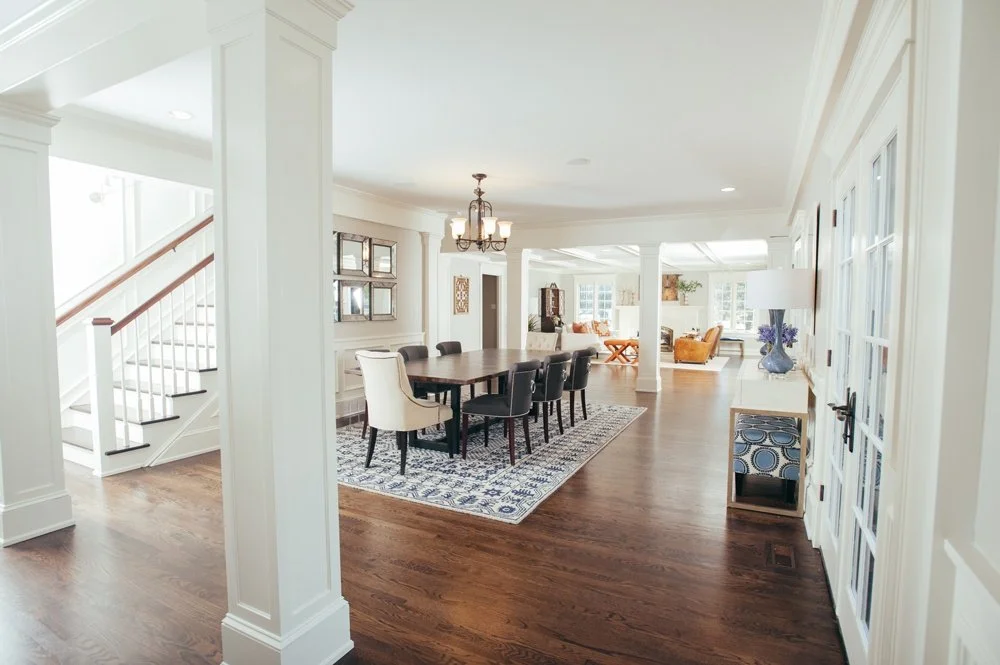 Spacious, well-lit dining and living area with dark hardwood floors, white walls, a chandelier over the dining table, and large windows in the background.