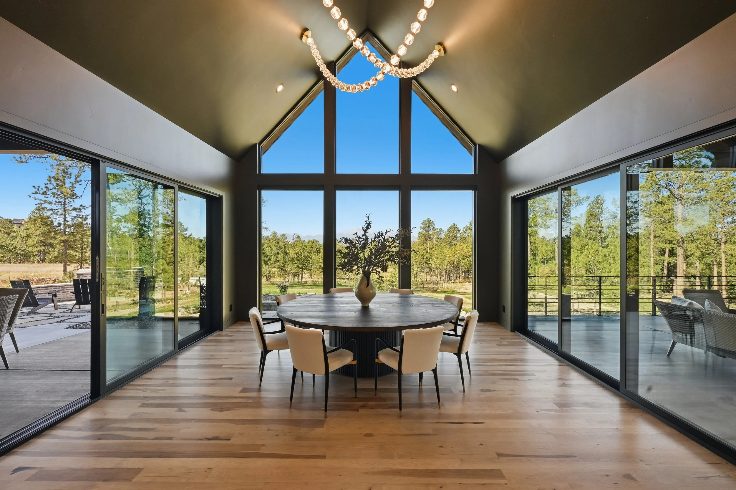 Modern dining room with a round table and eight beige chairs, large windows showing trees and blue sky, and a chandelier hanging from a high, vaulted ceiling.