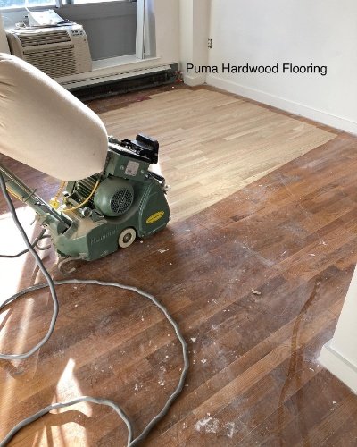 A room with two types of hardwood flooring, one being replaced by Puma hardwood flooring, with sanding and polishing equipment visible.