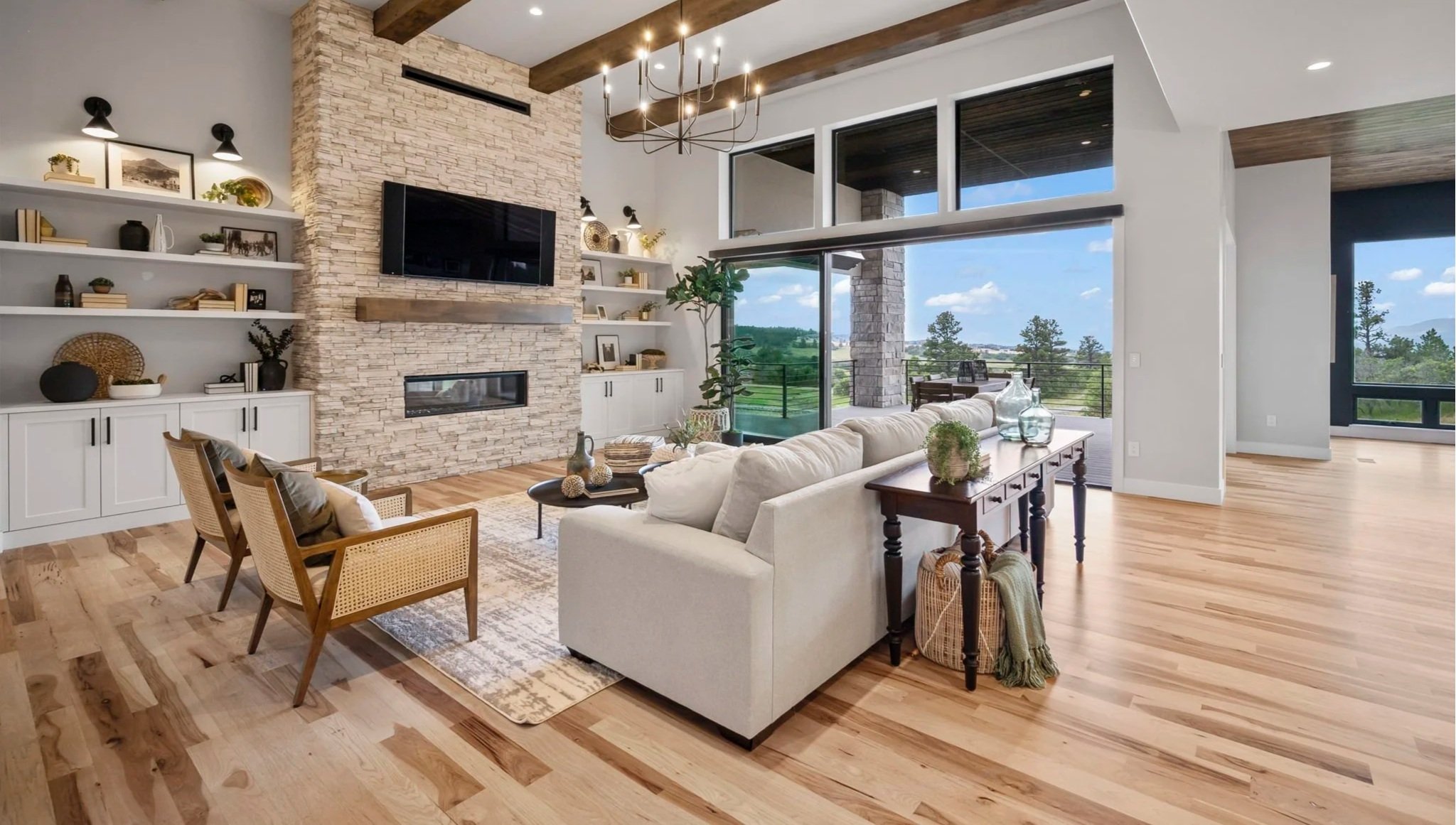 Open living room with large windows showing green landscape, stone fireplace, TV, white sofa, wooden chairs, and decorative items like vases and plants.