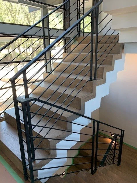 Interior view of a modern staircase with wooden steps and black metal railings, leading from a lower floor to an upper floor, with large windows and trees visible outside.