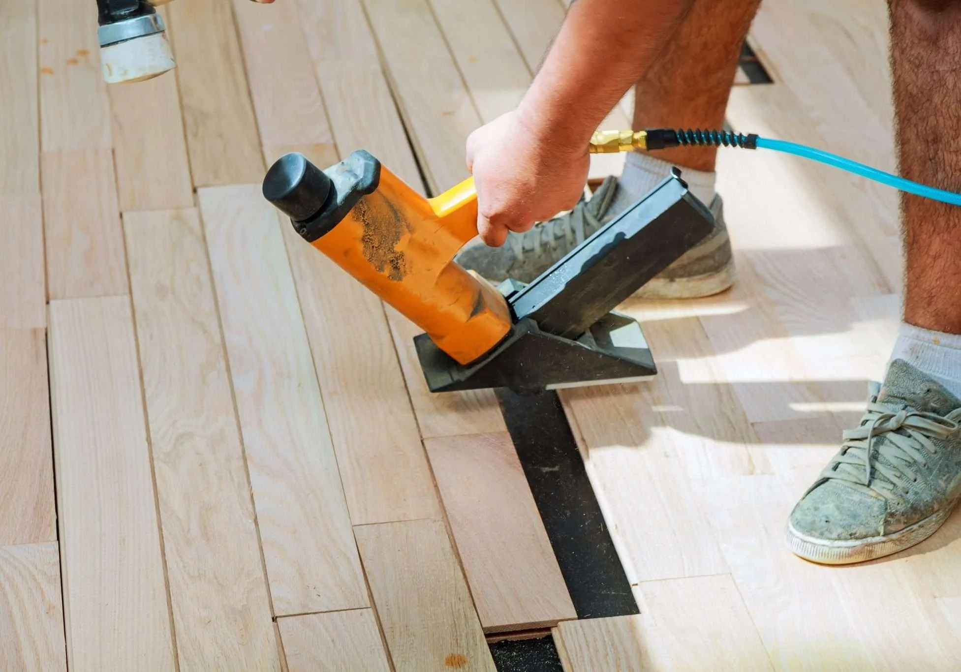 Person installing hardwood flooring, kneeling on the floor, holding a pneumatic nail gun.