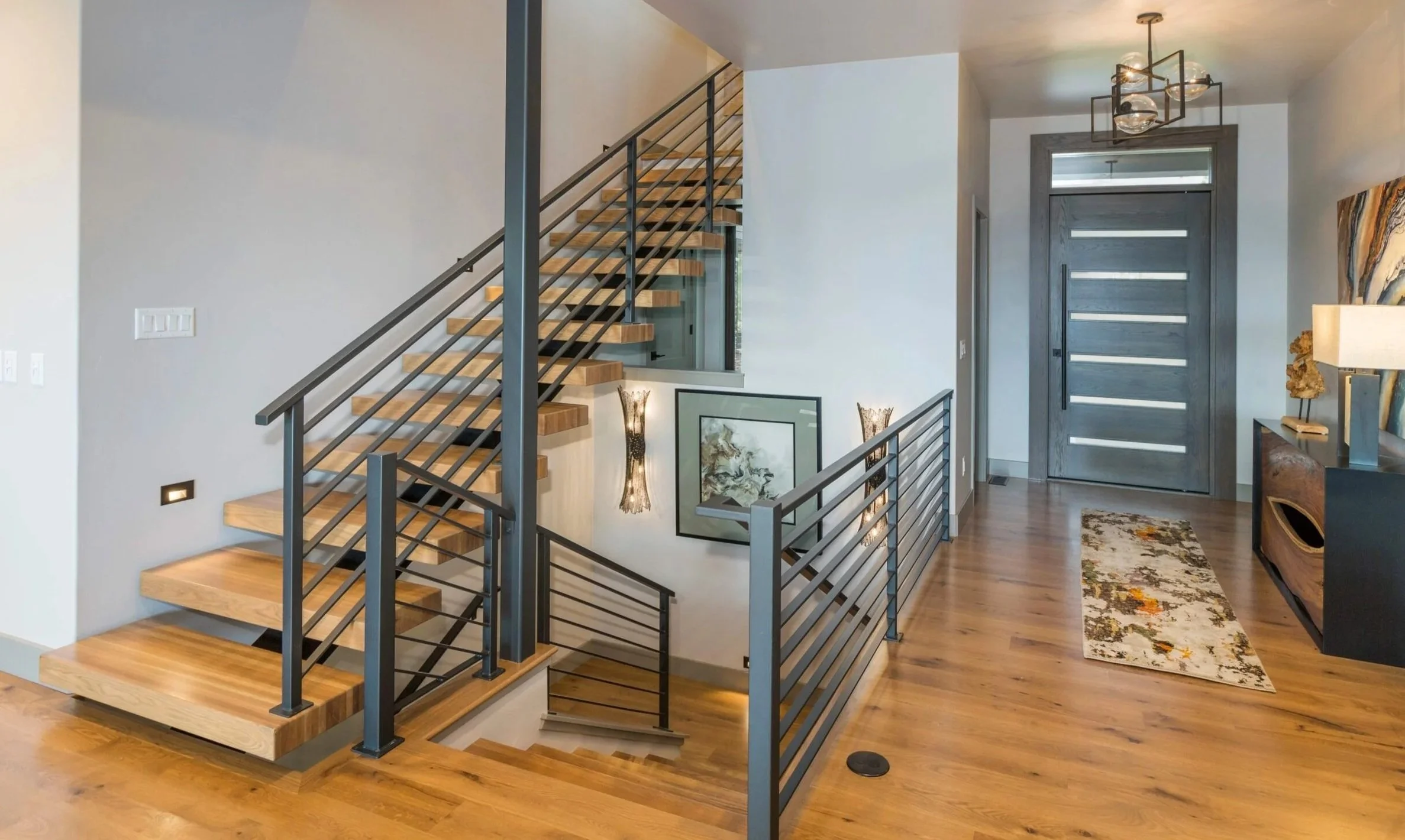 Modern interior entryway with a wooden staircase and metal railing, dark gray front door with horizontal glass panels, hardwood flooring, decorative artwork, rug, and lighting fixtures.