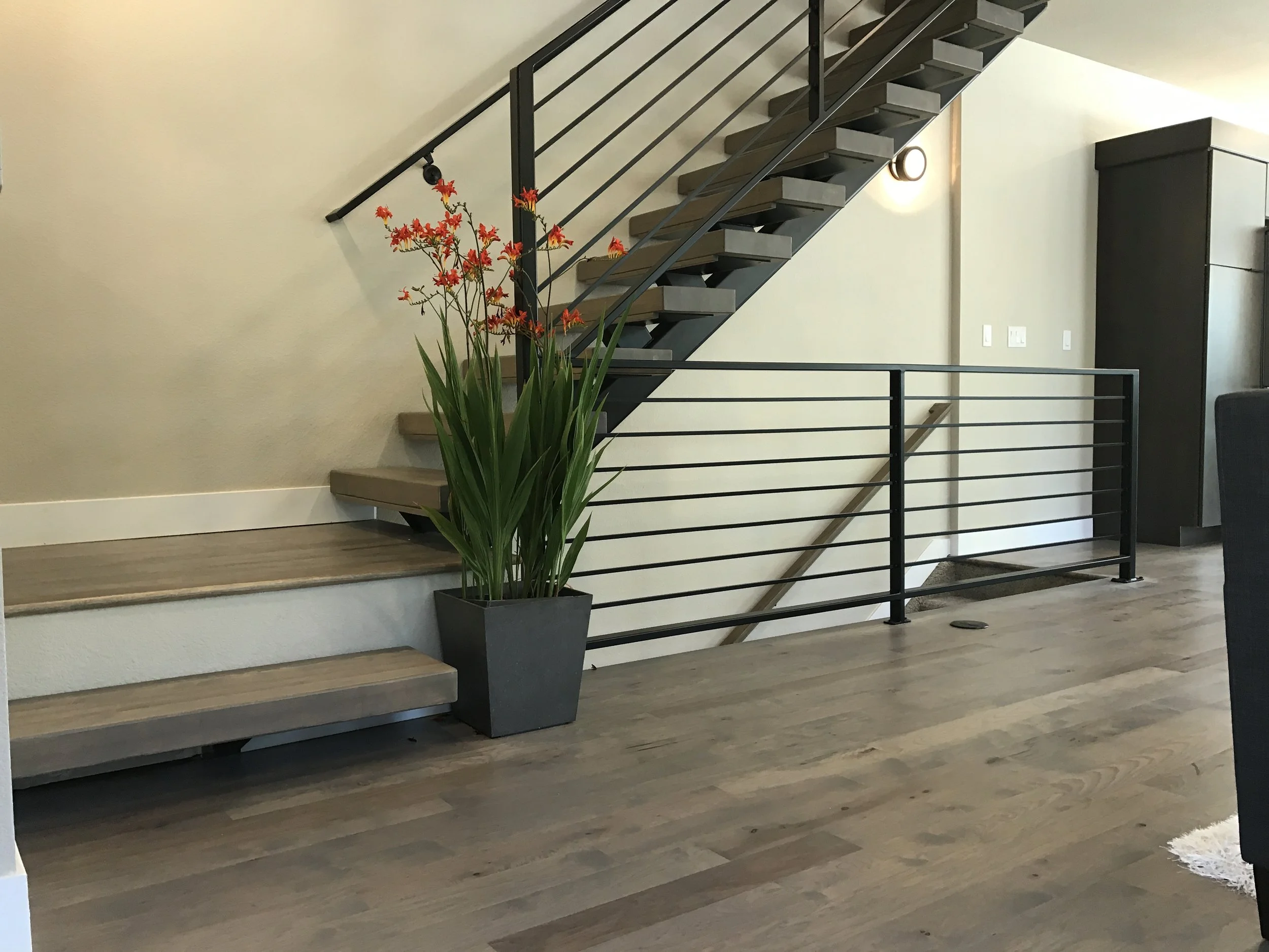 Interior view of a modern staircase with wooden steps, black metal railing, and a potted plant with red flowers next to the stairs. There is a black cabinet against the wall and a wall light fixture nearby.