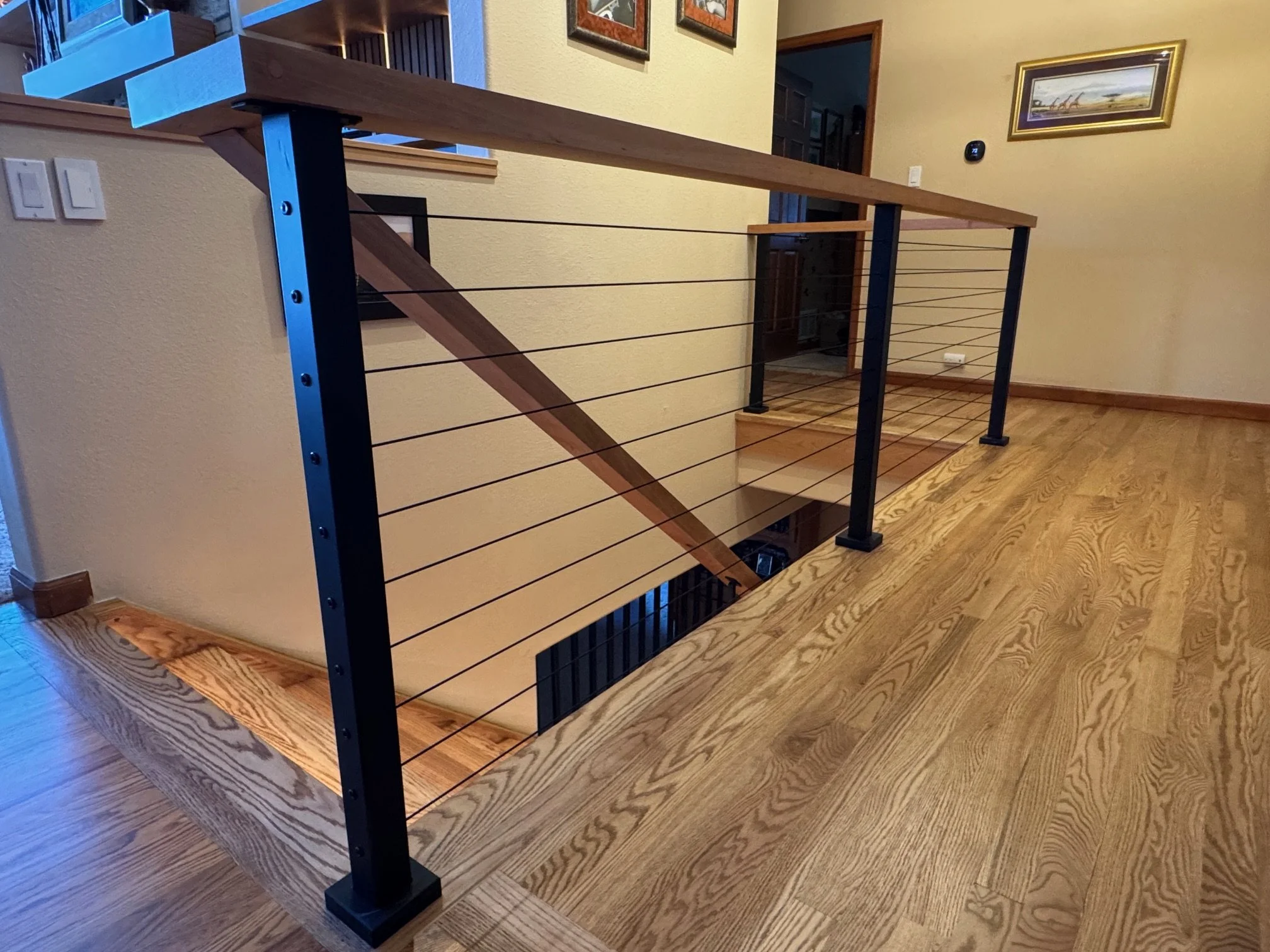 Interior view of a staircase railing made of black metal posts, wooden handrail, and horizontal cables, in a home with hardwood floors, beige walls, and framed artwork.