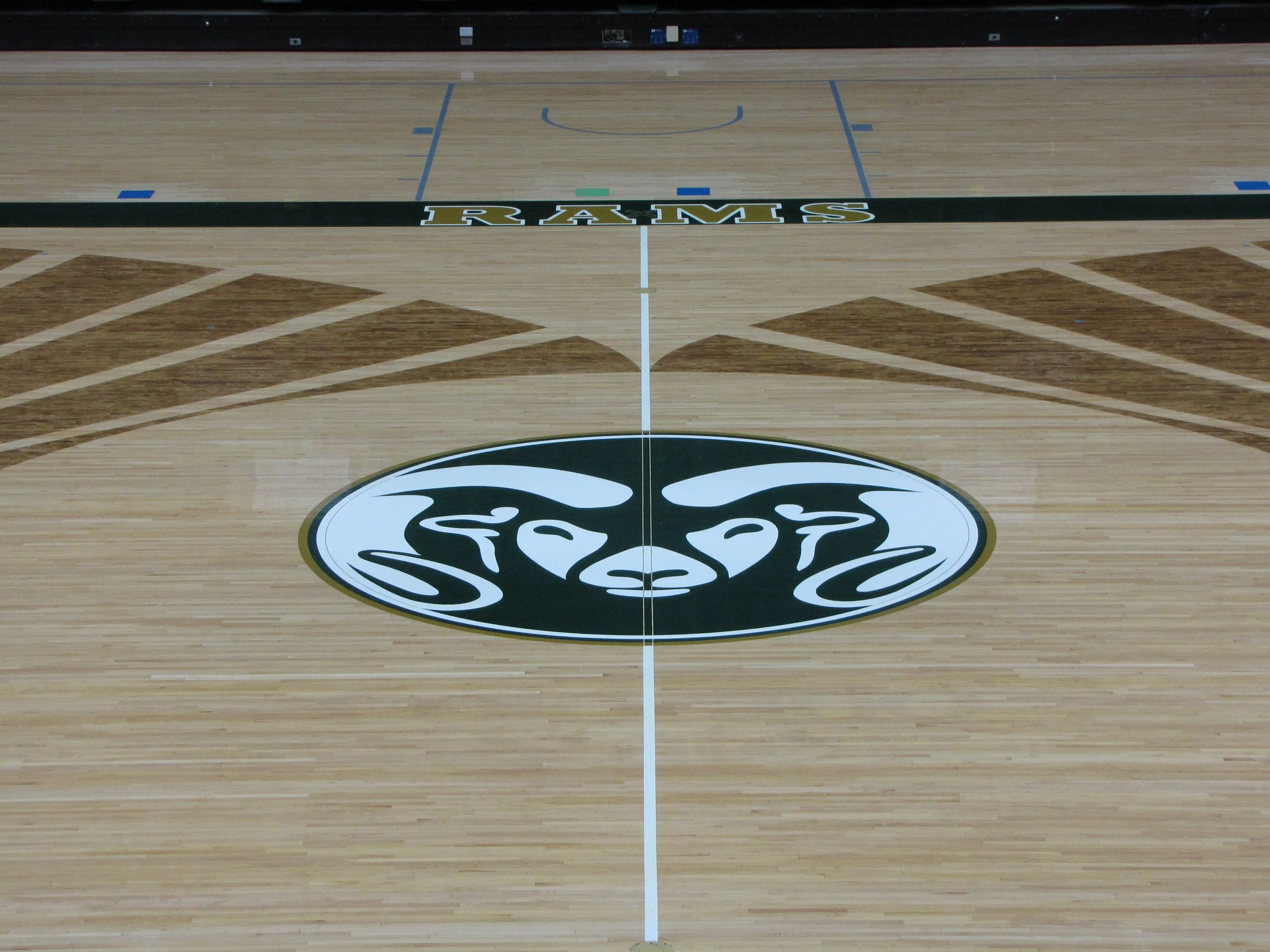 An empty basketball court with a large Rams logo at the center, hardwood flooring, and colorful lines marking the court.