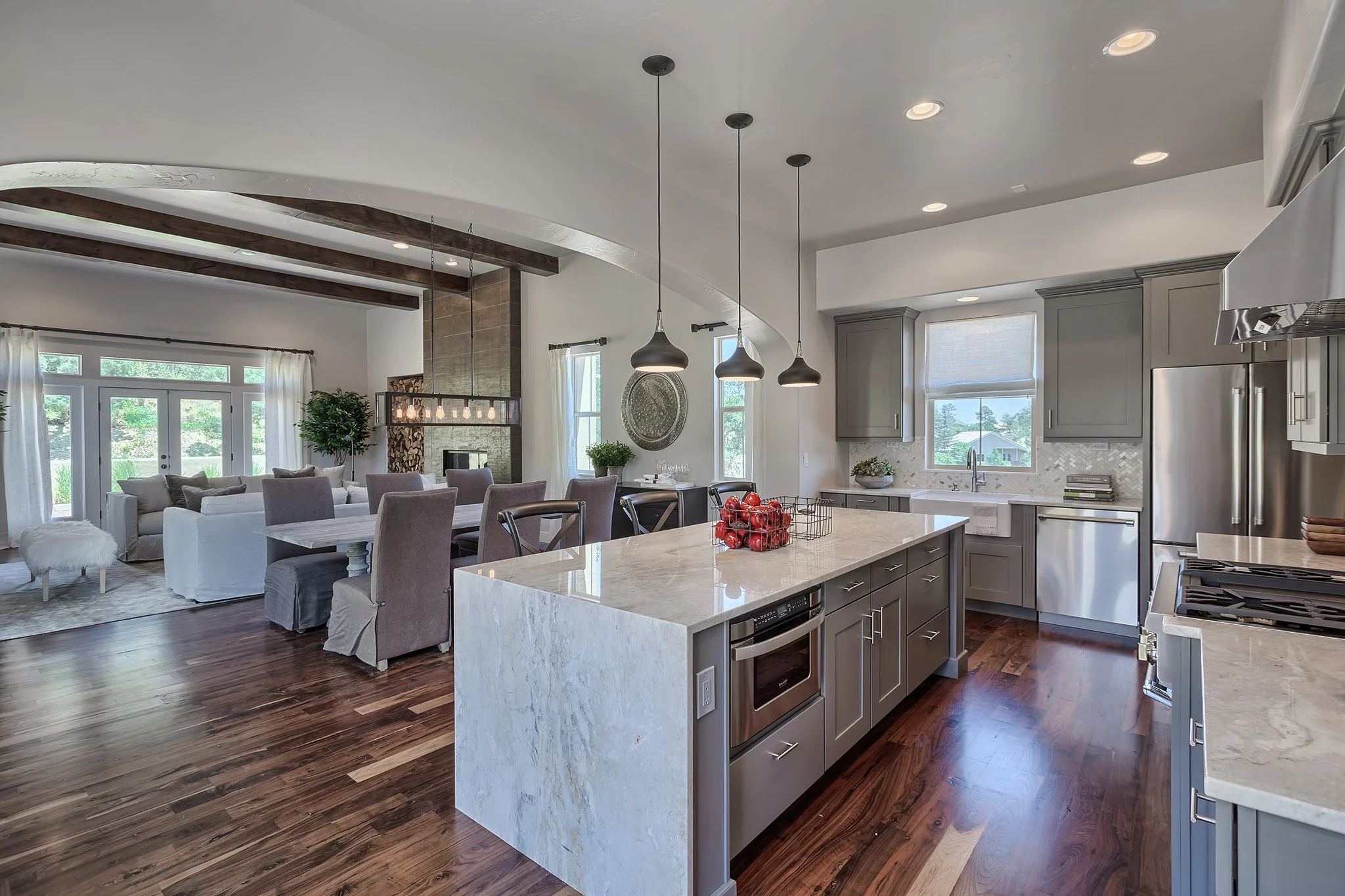 Open-concept kitchen and living room with hardwood floors, gray cabinets, white countertops, and large windows with curtains. The kitchen island has a basket of red apples and pendant lights above. The living area has a white sofa, gray chairs, and a fireplace with a wood decor wall.
