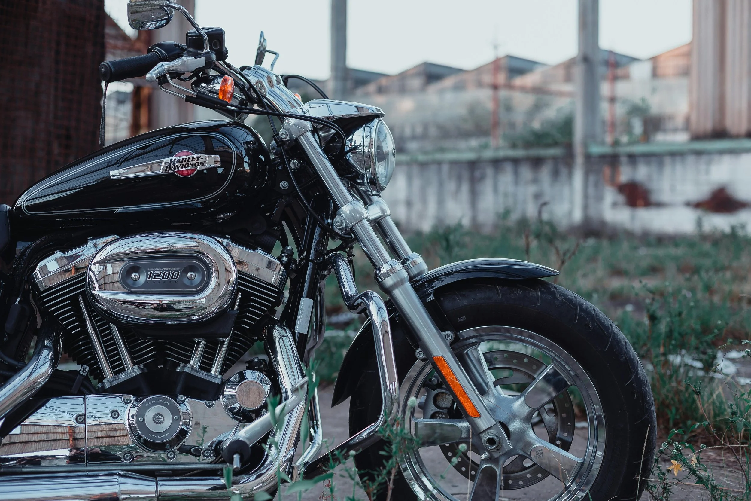Close-up of a black Harley-Davidson motorcycle parked outdoors on grass with an industrial background of brick buildings and a metal fence.