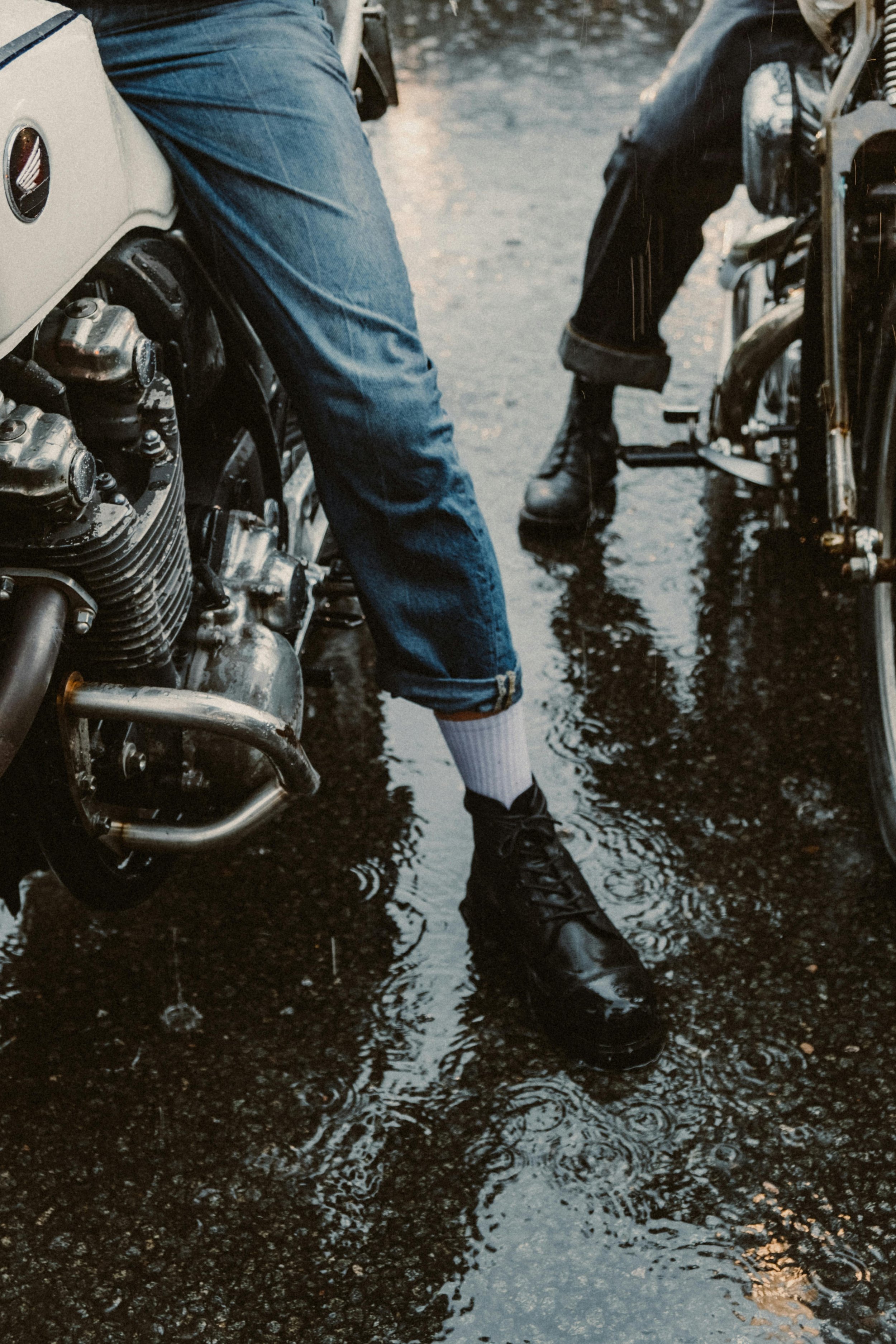 People sitting on motorcycles with water on the ground and reflection of their legs.