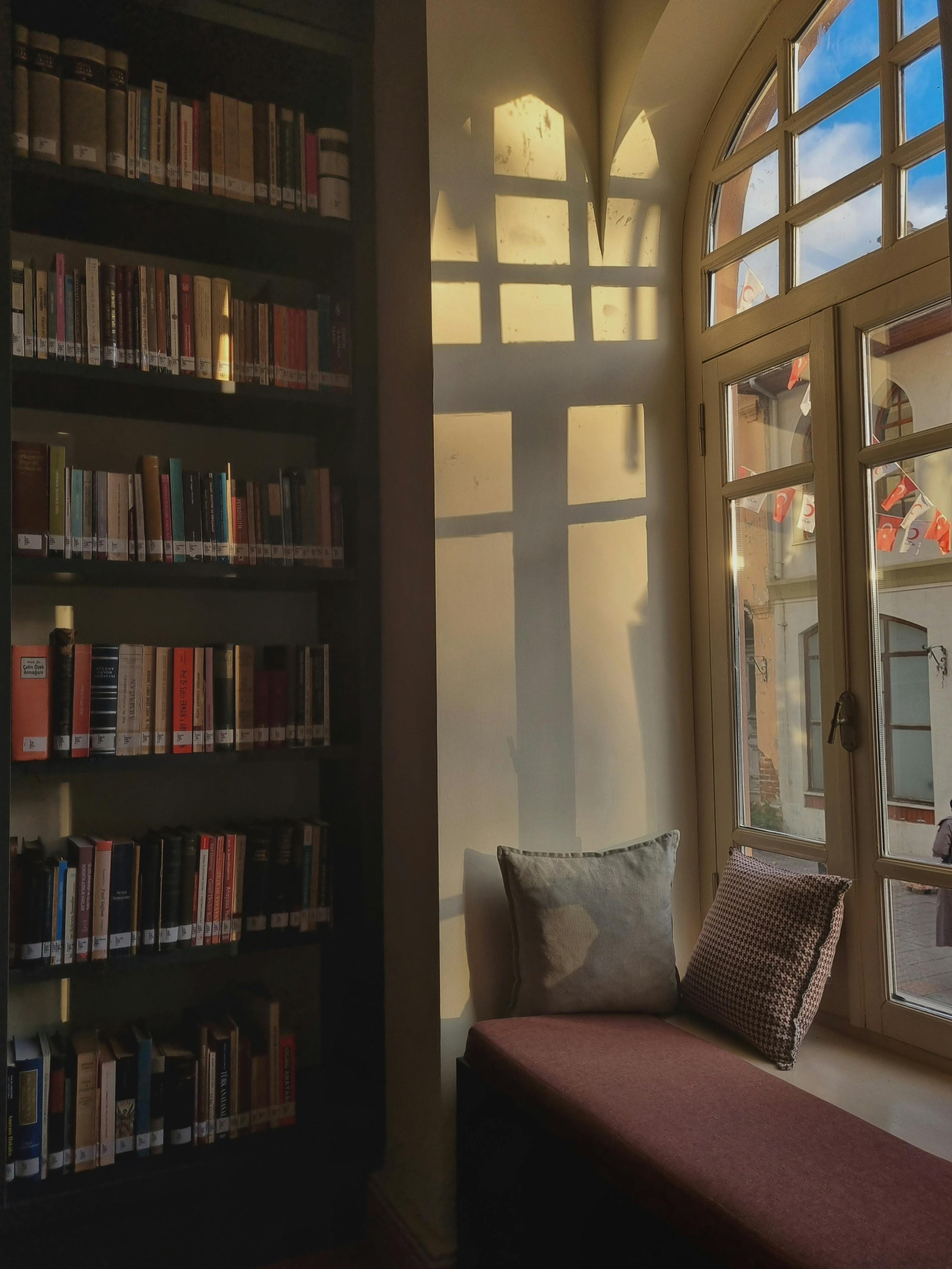 Sunlight creates shadows of window panes on a white wall in a library corner. A black bookshelf filled with books is on the left, and a cushioned window seat with pillows is on the right.