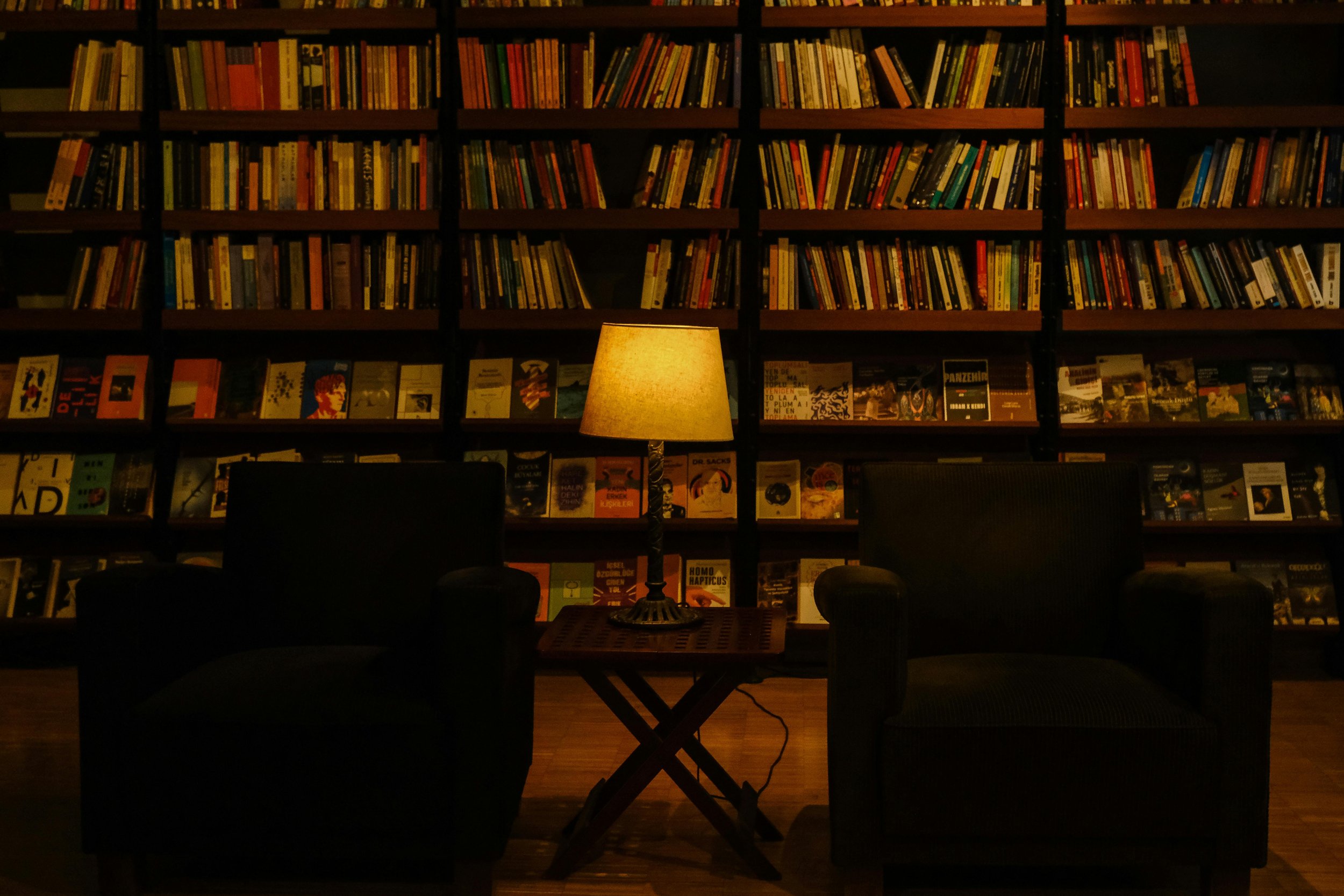 A cozy library with dark wood bookshelves filled with colorful books, a table with a lamp casting warm light, and black chairs arranged around the table.