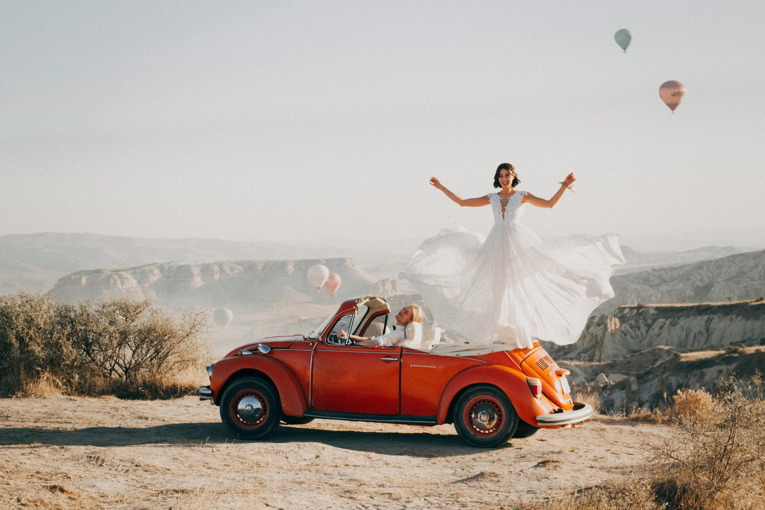 A woman in a flowing white dress standing on top of a red vintage convertible car with a man sitting inside. The scene is set in a desert landscape with hot air balloons floating in the sky.