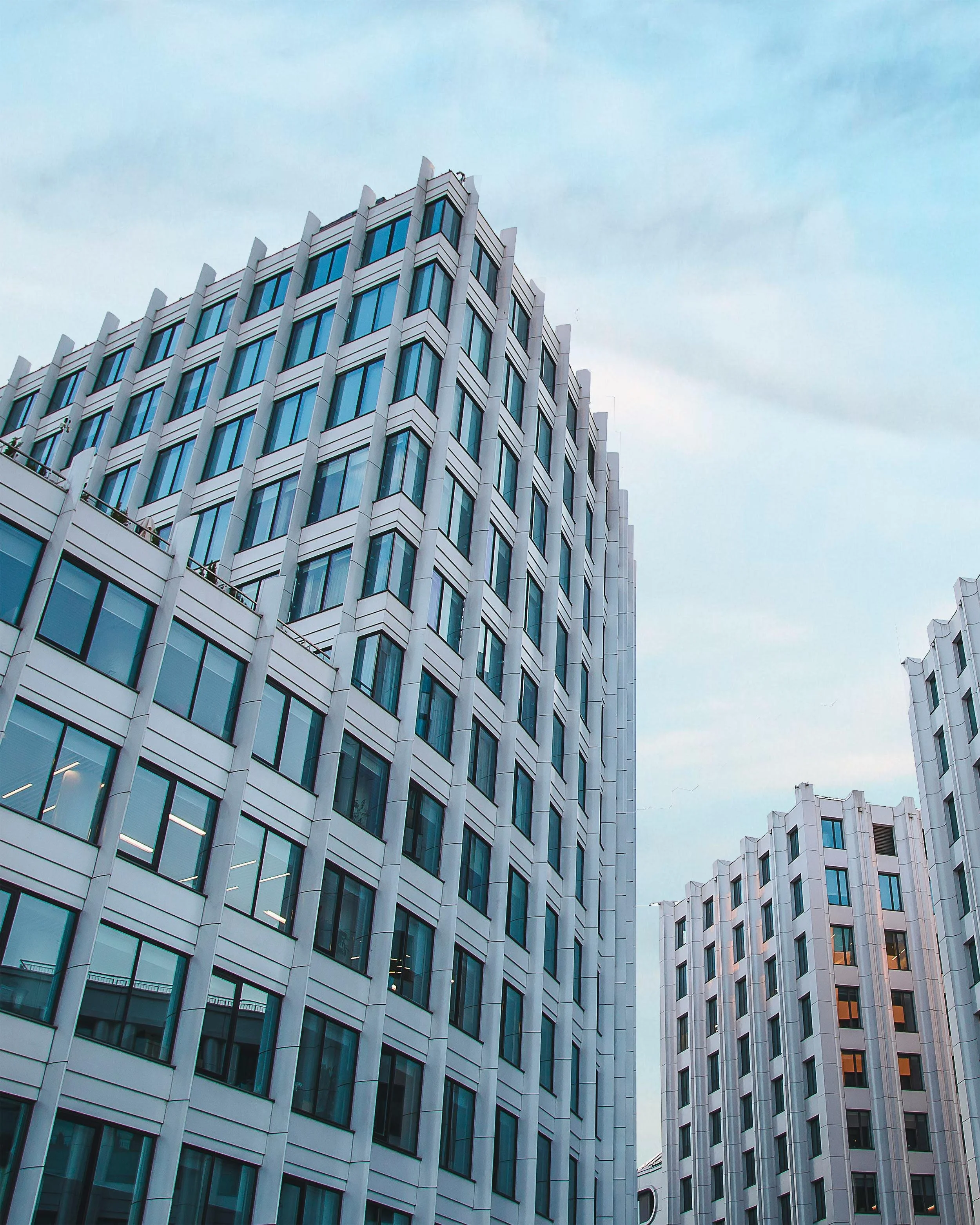 A view of modern white office buildings with many windows, some with lights on, under a blue-gray sky.
