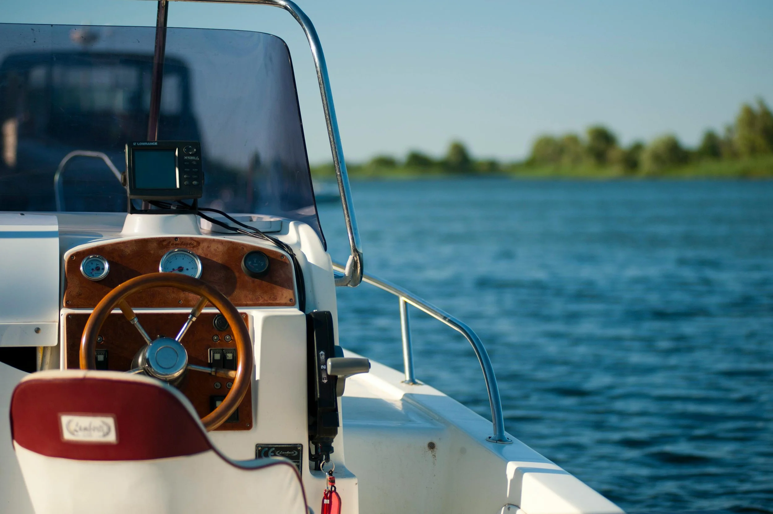 View of a boat's dashboard with a steering wheel, gauges, and electronic navigation device on lake with land and trees in the background.
