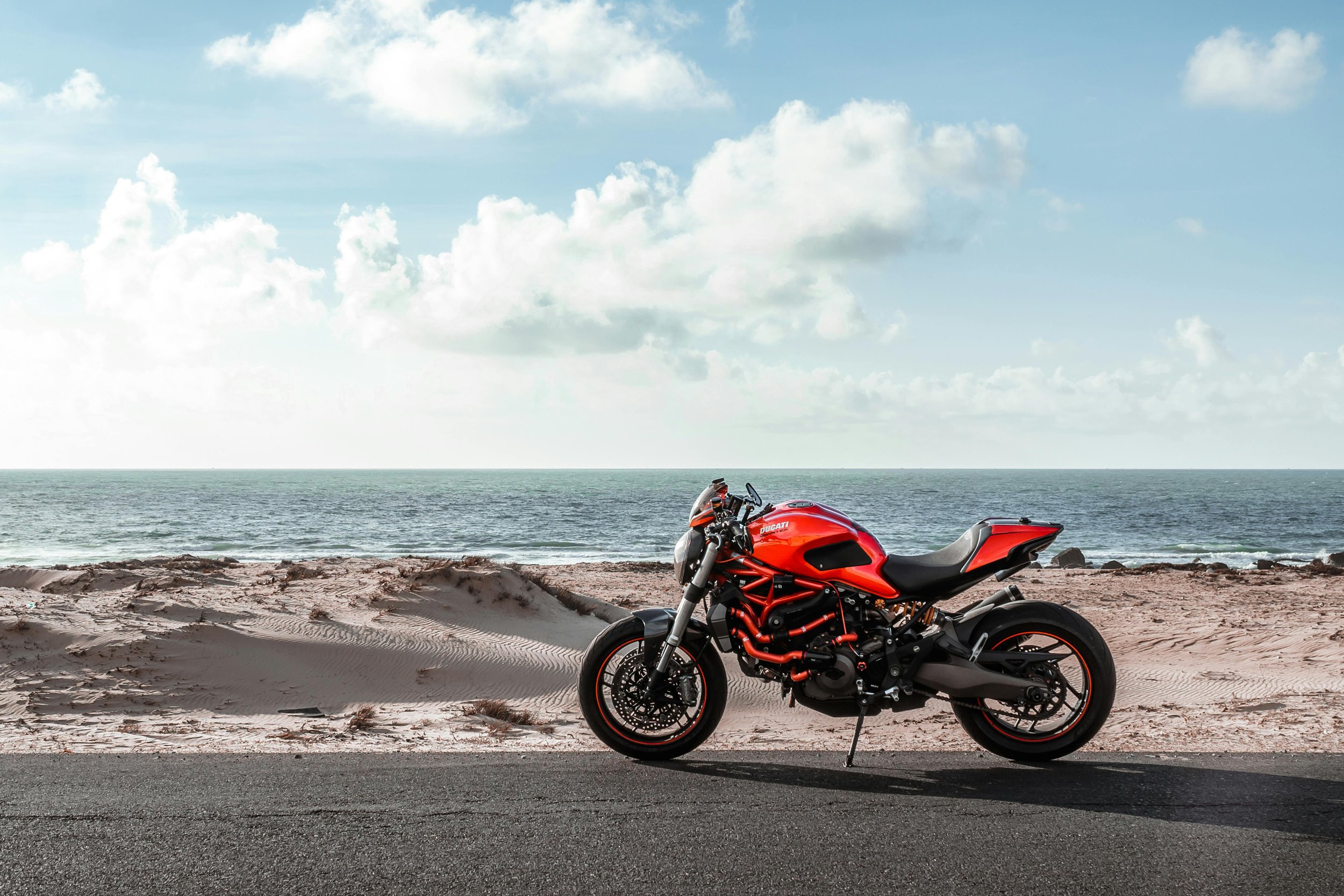 A red Ducati motorcycle parked on the side of a road near a beach, with sand, rocks, the ocean, and a partly cloudy sky in the background.
